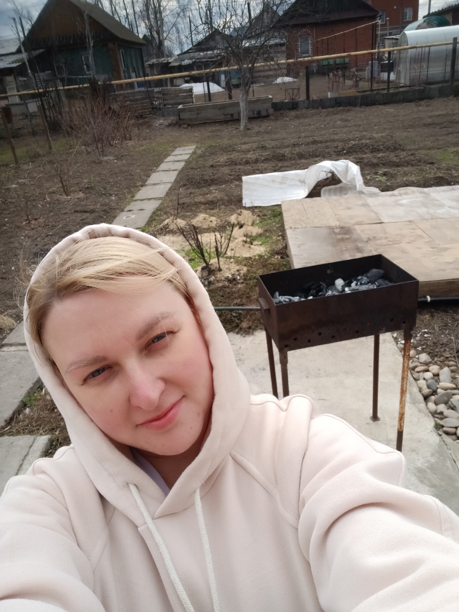 A blonde individual, likely female, wearing a light beige or off-white hooded sweatshirt, takes a selfie in what appears to be a backyard or garden in Chaikovsky, Russia. She looks directly at the camera with a calm, subtle smile.
The setting is an outdoor area with a mix of concrete paving and bare earth, suggesting early spring or late autumn due to the lack of green foliage and the presence of bare trees in the background. To the right of the person, a dark metal brazier (mangal) filled with charcoal stands on short legs on a paved surface, indicating preparation for grilling. A path made of concrete stepping stones extends from the middle ground into the background.
In the background, several houses are visible, including older wooden structures and a red brick house, along with fences and sparsely vegetated ground. A white tarp-like covering lies on the ground further back, and a greenhouse can be seen in the distance near the brick house. The sky is overcast, providing diffuse lighting. No text is visible in the image.