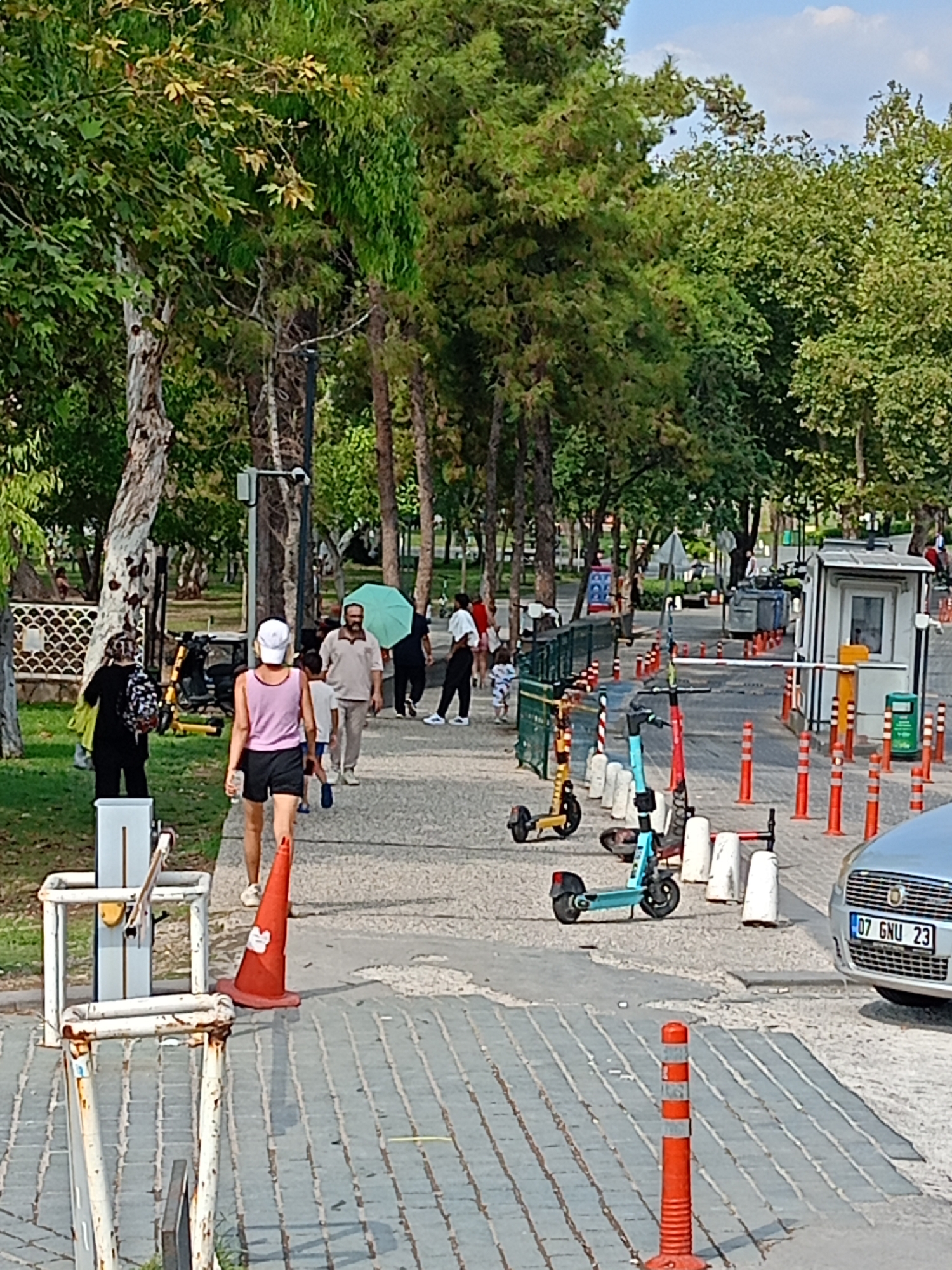 In Antalya, Turkey, a paved walkway in a park area is populated by several individuals and objects. On the left, two women are walking away from the camera. The woman in front has a backpack, and the woman behind her wears a pink tank top and black shorts. A bright orange traffic cone is placed on the walkway near them. Further down the path, a man in a beige polo shirt is walking with a group of children, one of whom is holding a light green umbrella.
To the right of the walkway, several electric scooters are parked in a row, including yellow, blue, and red models. A security booth with a white exterior and a yellow barrier arm is visible, along with numerous orange and white bollards that delineate a path. A light silver car with the license plate "07 GNU 23" is parked on the right side of the scene. The environment is characterized by lush green trees lining the walkway, suggesting a park or recreational area. The sky above is a pale blue with scattered clouds.