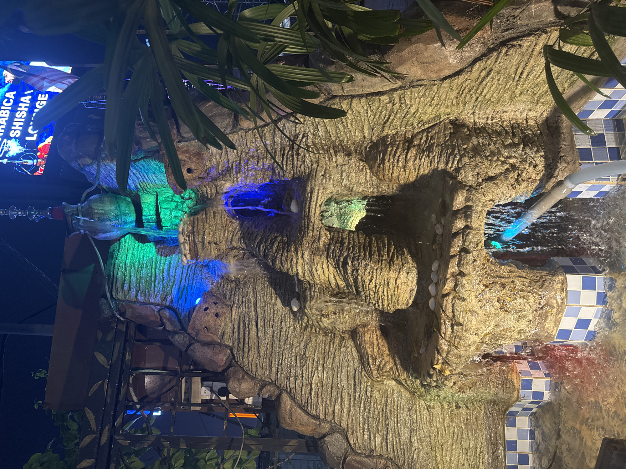 The image displays an illuminated artificial rock waterfall at night, likely situated at a commercial establishment in Kuala Lumpur, Malaysia. The central feature is a multi-tiered rock formation, intricately textured to resemble natural stone, with water cascading down its various levels. The waterfall is vibrantly lit by colorful artificial lights, casting prominent green and blue glows onto the flowing water and the rock surfaces, creating a dynamic and eye-catching display.
In the upper left portion, large green plant leaves hang over the scene, partially obscuring a bright blue neon sign. The visible text on this sign clearly reads "ARABICA SHISHA LOUNGE," indicating the type of venue. Below the foliage and near the sign, a glass shisha pipe is discernible. Water collects or flows into a basin on the right side of the frame, which is adorned with distinctive blue and white checkered tiles. A gray pipe, part of the water circulation system, is also visible. Electrical outlets and wiring are noticeable on a support structure towards the bottom left, revealing the technical setup behind the artificial feature. The dark background emphasizes the nighttime setting, with all illumination originating from the artificial lighting, contributing to a vibrant and atmospheric ambiance for patrons of the lounge.