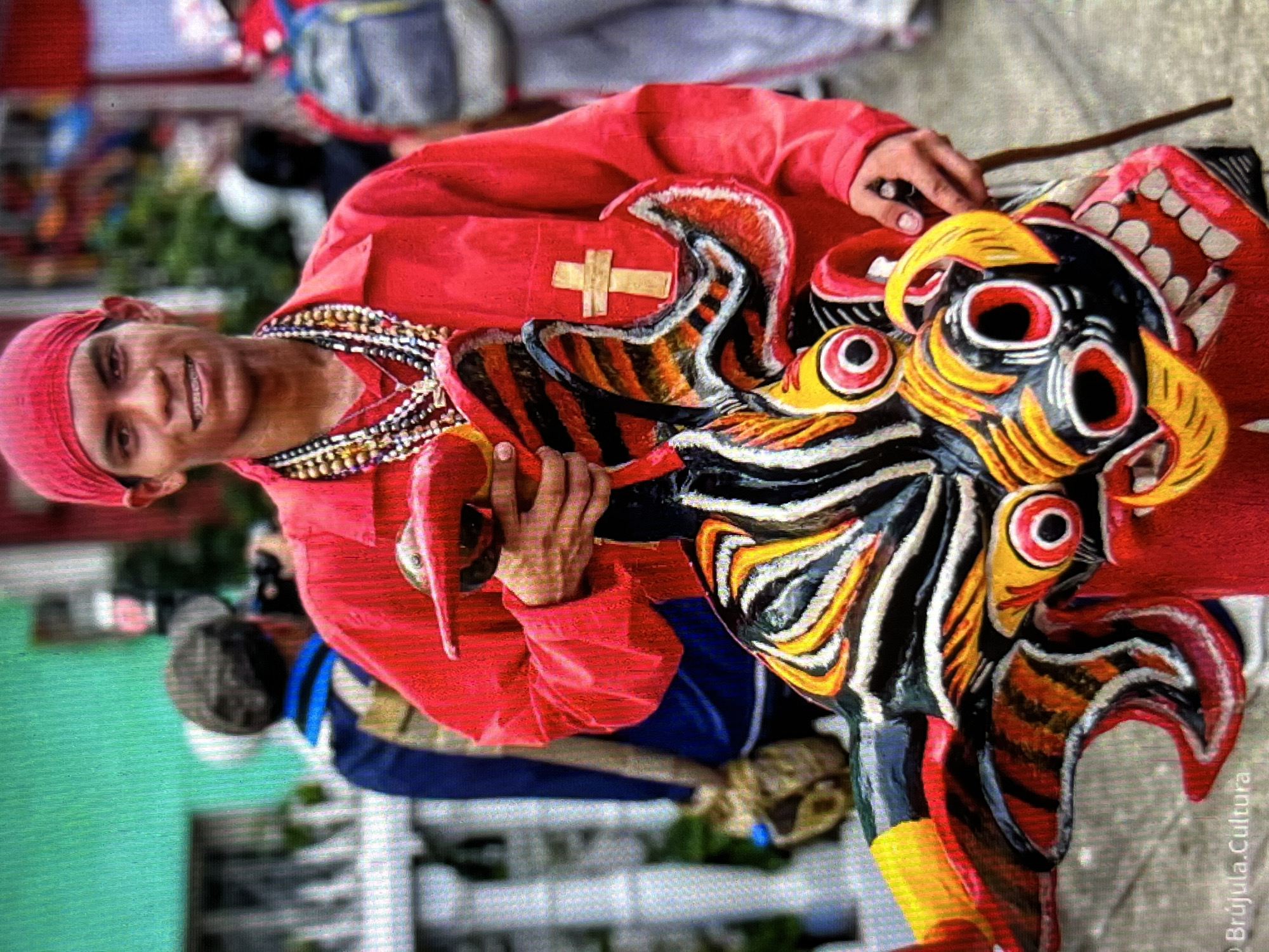 A young man in a red head covering and shirt, adorned with multiple beaded necklaces, stands in front of a white fence. He holds a colorful, multi-faceted mask with a red, yellow, black, and white striped pattern. The mask has large, round, red-rimmed eyes with black pupils and a wide mouth with white teeth. A small, red object, possibly a maraca or rattle, is held in his left hand. A wooden stick is held in his right hand, to the right of the mask. In the background, other people are visible, including one wearing a blue jacket and another with a backpack. The overall scene suggests a cultural event or festival. The location is identified as Tinaquillo, Venezuela.