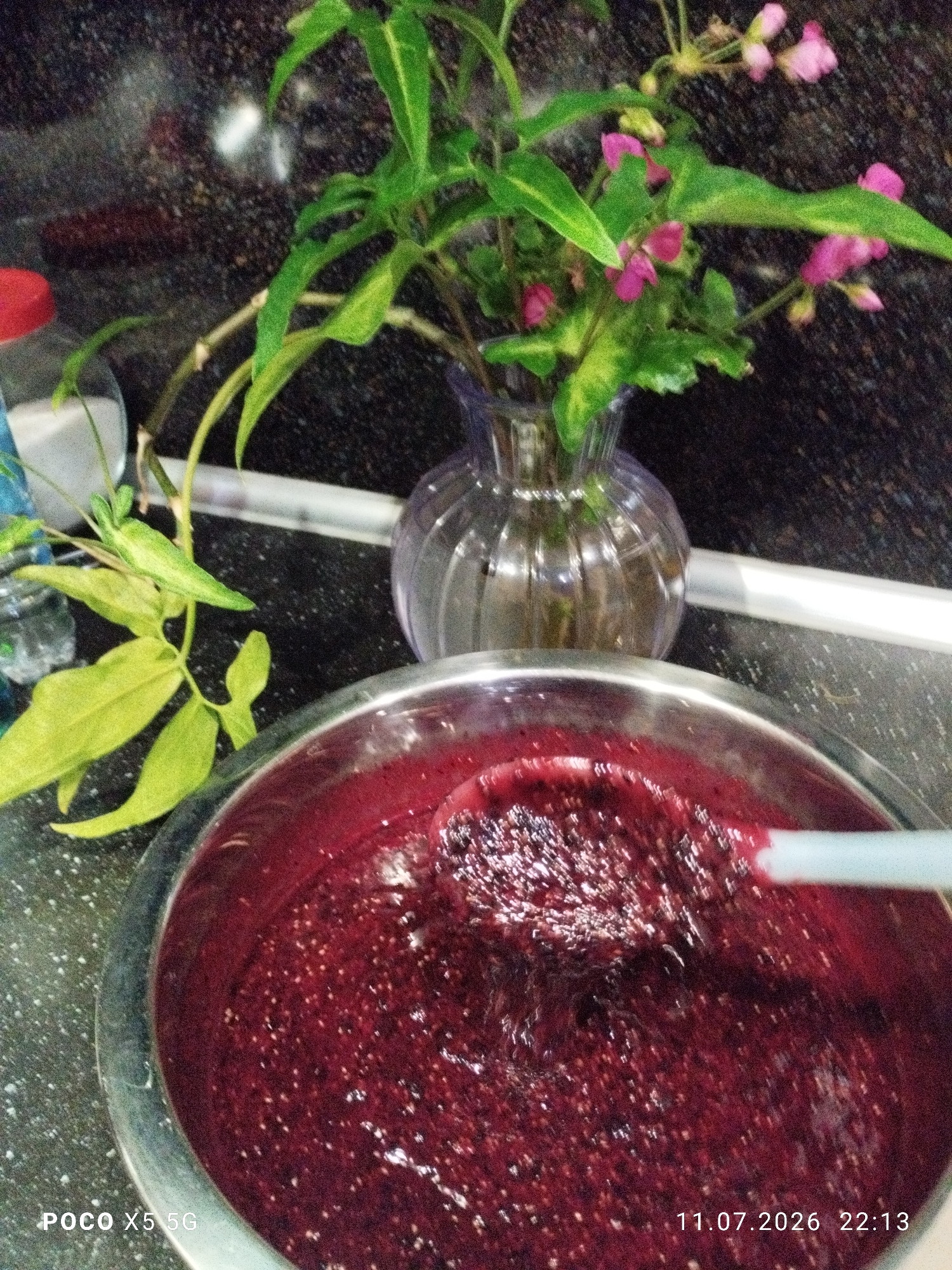 A close-up, high-angle shot shows a metallic bowl filled with a deep red, possibly berry-based, liquid. A white stirring utensil is partially submerged in the liquid. To the left and behind the bowl, a plant with green leaves and small pink flowers in a clear, ribbed vase is visible. A red-capped bottle and a clear glass are also on the counter to the left of the bowl. The countertop is dark and speckled. The bottom left corner of the image displays "POCO X5 5G" and the bottom right corner shows "11.07.2026 22:13". The scene appears to be indoors, likely a kitchen, as suggested by the countertop and the presence of food preparation items. There is no indication of the city of Brek, Russia in the image.