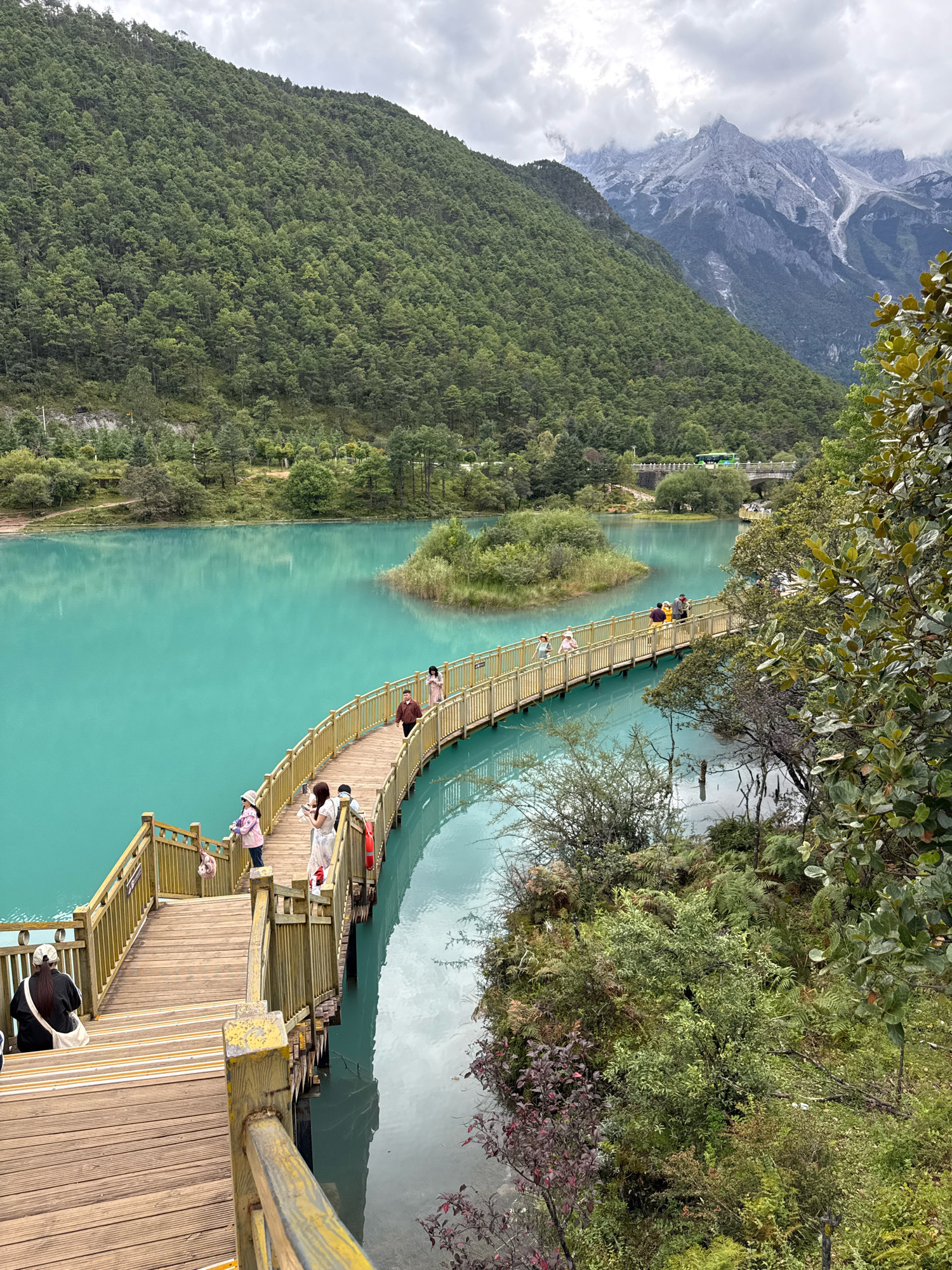 The image depicts a picturesque landscape in Yulong Nakhi Autonomous County, China. A vibrant turquoise lake dominates the scene, bordered by lush green mountains. A wooden boardwalk winds along the edge of the lake, with several tourists strolling on it. Some people are taking photos, while others are simply enjoying the view. The boardwalk is elevated, offering a clear perspective of the water and the surrounding scenery. In the background, snow-capped mountains pierce the cloudy sky, adding a dramatic touch to the serene environment. The vegetation is dense and varied, with a mix of trees and shrubs contributing to the natural beauty of the location. The overall atmosphere is one of tranquility and natural wonder.