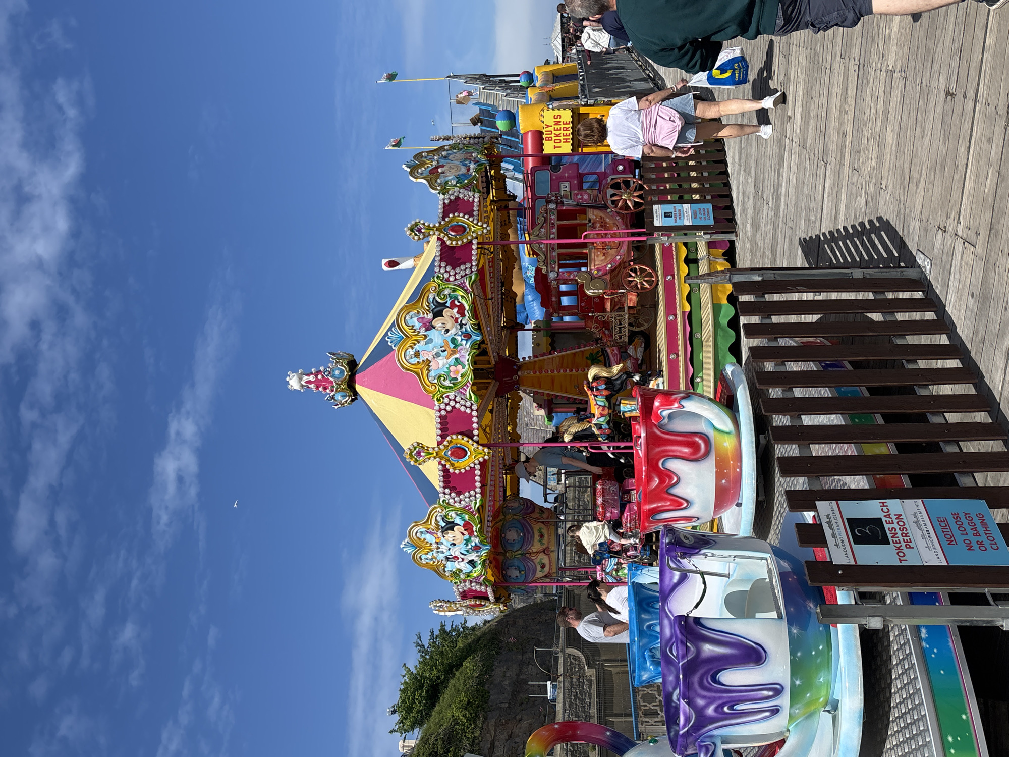 This image shows a carousel on Llandudno Pier in the United Kingdom. The carousel is decorated with colorful patterns and cartoon characters and is surrounded by a wooden platform. People are standing on the platform, some of them are on the carousel, and there is a sign indicating that tickets are required to ride. The sky is blue with a few clouds. The sign says, "3 tokens each person" and "no loose or baggy clothing."