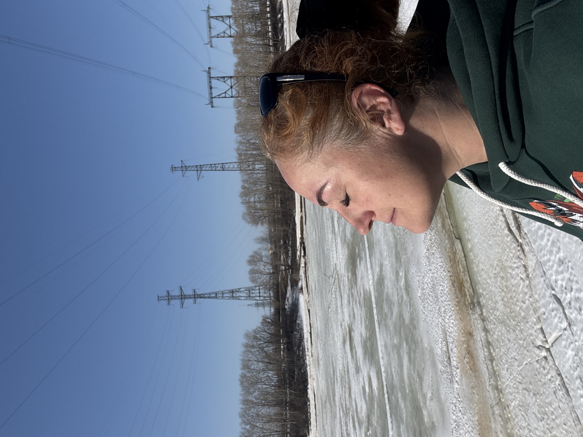 The image shows a person with reddish hair, wearing sunglasses on their head and a dark green hooded sweatshirt, standing beside a frozen body of water, possibly a river or lake, in Chaikovsky, Russia. The frozen surface appears cracked and textured. In the background, there are trees without leaves and power line towers stretching into the blue sky.