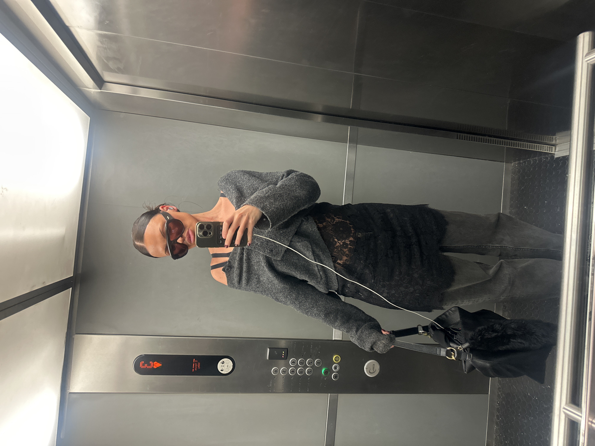 A person, appearing to be a young adult with hair pulled back, is captured taking a mirror selfie inside an elevator with reflective metallic walls and a dark floor. The individual is wearing dark sunglasses that, along with a smartphone held up, obscure most of their face. Their attire includes a dark, possibly lace or sheer, camisole or top worn under a loose-fitting, textured grey knitted cardigan or sweater with an off-shoulder style. Dark-wash trousers or jeans complete the casual ensemble. An earphone wire hangs from the phone. In one hand, they hold a black handbag featuring gold-toned hardware and a fluffy black attachment.
The brushed metal interior of the elevator reflects ambient light. The control panel features a digital display showing "E3" with an upward arrow, indicating the elevator's current status or destination. Visible text on the panel includes "GOODS ONLY," "MAX 12 PERSONS," "1000 Kg," and the city name "London," confirming the location as London, United Kingdom. Below these details, floor selection buttons numbered 0 through 8 are present, along with door open/close buttons, an alarm bell, a phone icon, and a green illuminated button, likely signifying a selected floor or active function.