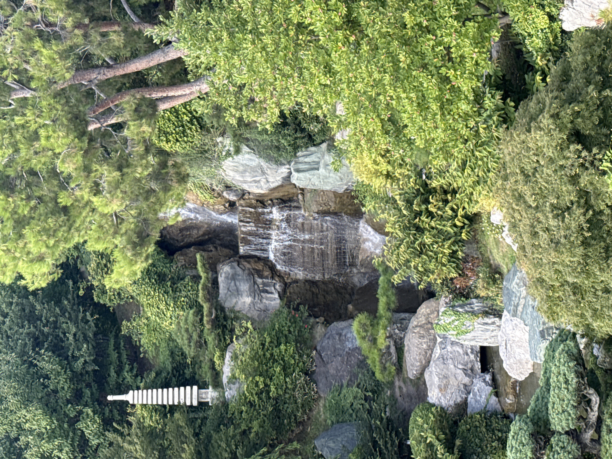This image depicts a meticulously landscaped Japanese garden, likely located within the Симеизский поселковый совет of Ukraine. The scene features a multi-tiered waterfall cascading over rocks into a tranquil pool below. To the left of the waterfall, a white, multi-tiered pagoda-like structure stands amidst lush greenery. The garden is characterized by a variety of trees, shrubs, and carefully placed rocks, creating a serene and naturalistic environment. The overall impression is one of peace and meticulous design, characteristic of traditional Japanese garden aesthetics. There are no people or animals visible in the image.