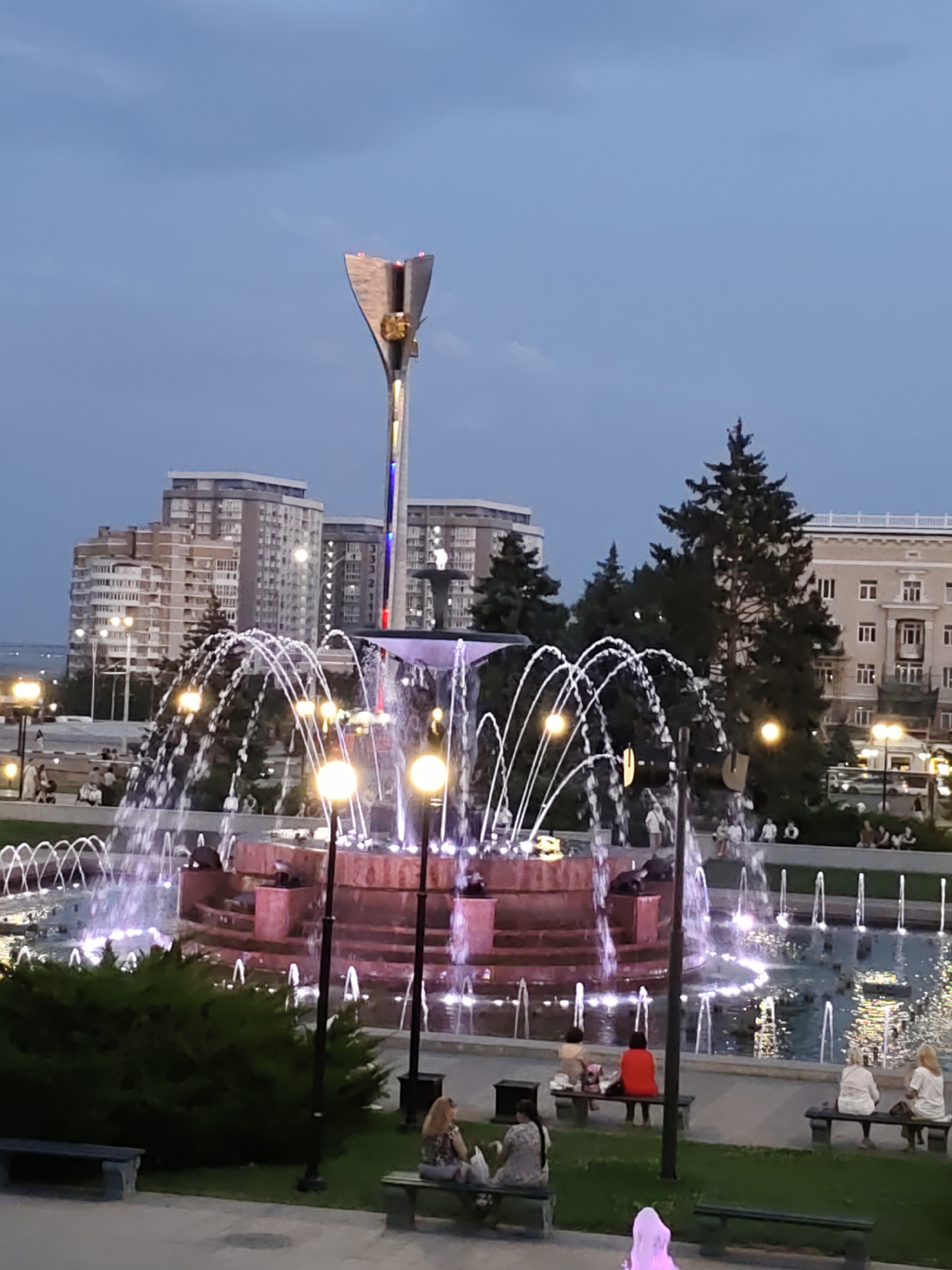 A fountain is illuminated with purple lights at dusk, with water jetting upwards in multiple streams. In the background, several apartment buildings and a spire-like monument are visible against a twilight sky. People are seated on benches and walking around the fountain area. The scene appears to be a public square or park.