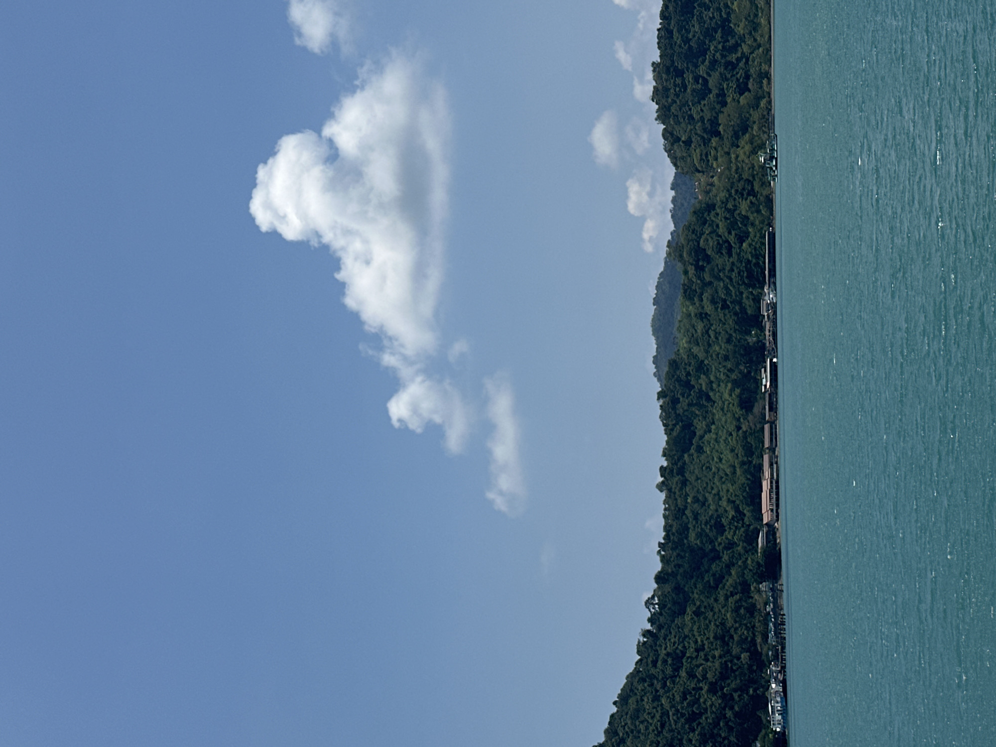 The image depicts a coastal scene in Baan Klong Son, Thailand. Lush, densely forested mountains extend along the coastline on the right, meeting the aquamarine water. Small buildings and structures are nestled amongst the trees. In the upper left quadrant, the blue sky contains fluffy white clouds.
