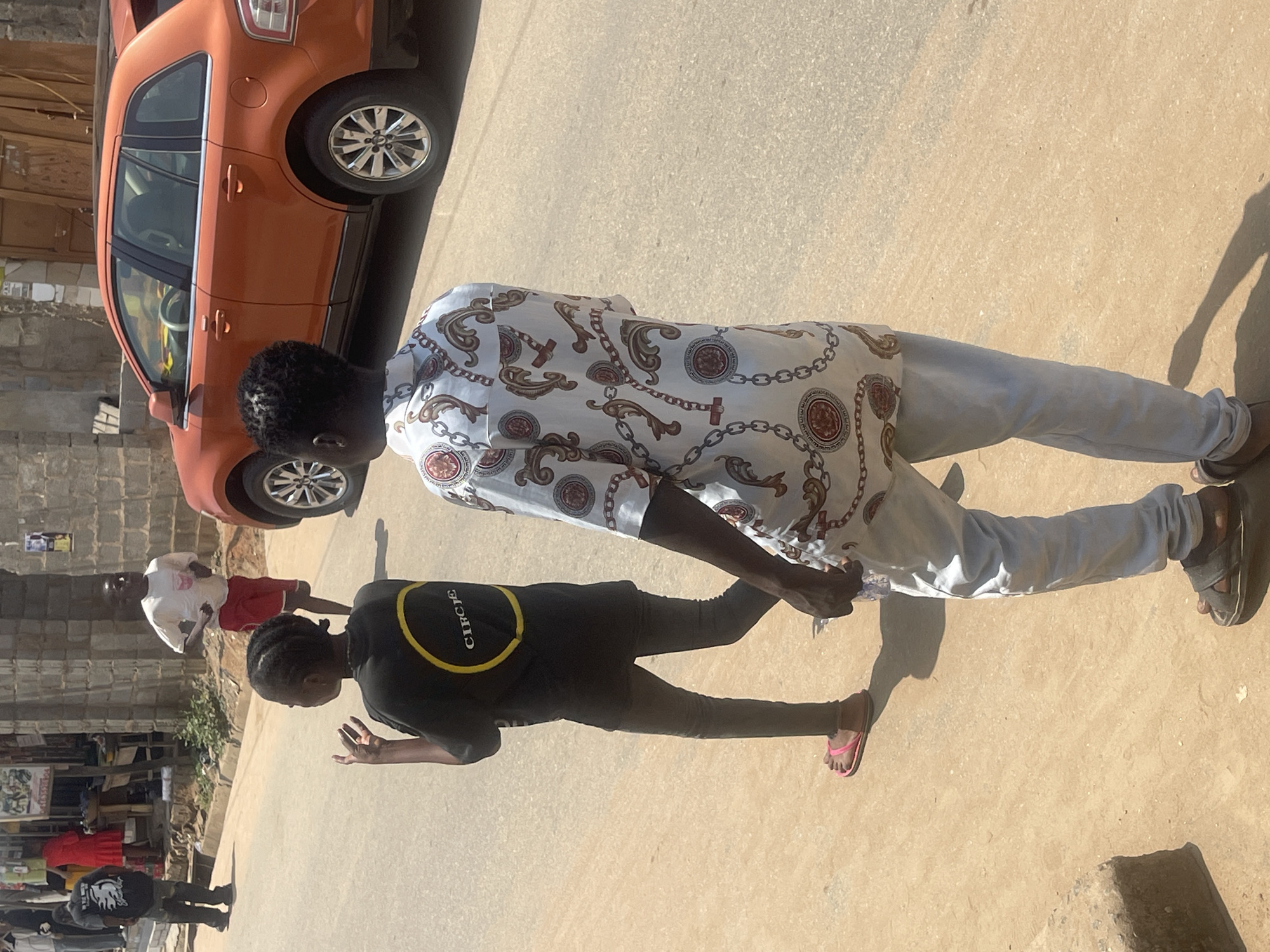 In Abuja, Nigeria, on a paved street, two young individuals walk casually. The person in the foreground, facing away from the camera, is wearing a white shirt with an intricate gold chain pattern and light blue trousers. They are also wearing dark sandals. To their left, a second individual, also facing away, wears a black t-shirt with a yellow circular logo that appears to read "CIRCLE," dark leggings, and pink flip-flops. Behind them, a boy in a white t-shirt and red shorts is visible running. An orange SUV is parked on the right side of the frame. The environment appears to be a commercial or residential area with buildings lining the street and vegetation in the background. The lighting suggests it is daytime.
