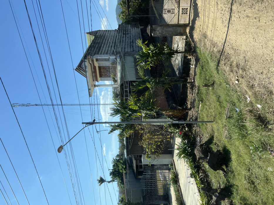 An outdoor scene features a tilted view of buildings, vegetation, and power lines. A white, two-story house with a porch and intricate detailing on the facade stands near palm trees. A communication tower rises above the power lines against a blue sky. Lower-lying houses and fences are visible in the distance. Patches of dirt, grass, and greenery border a walkway or road. A street lamp is also seen.
