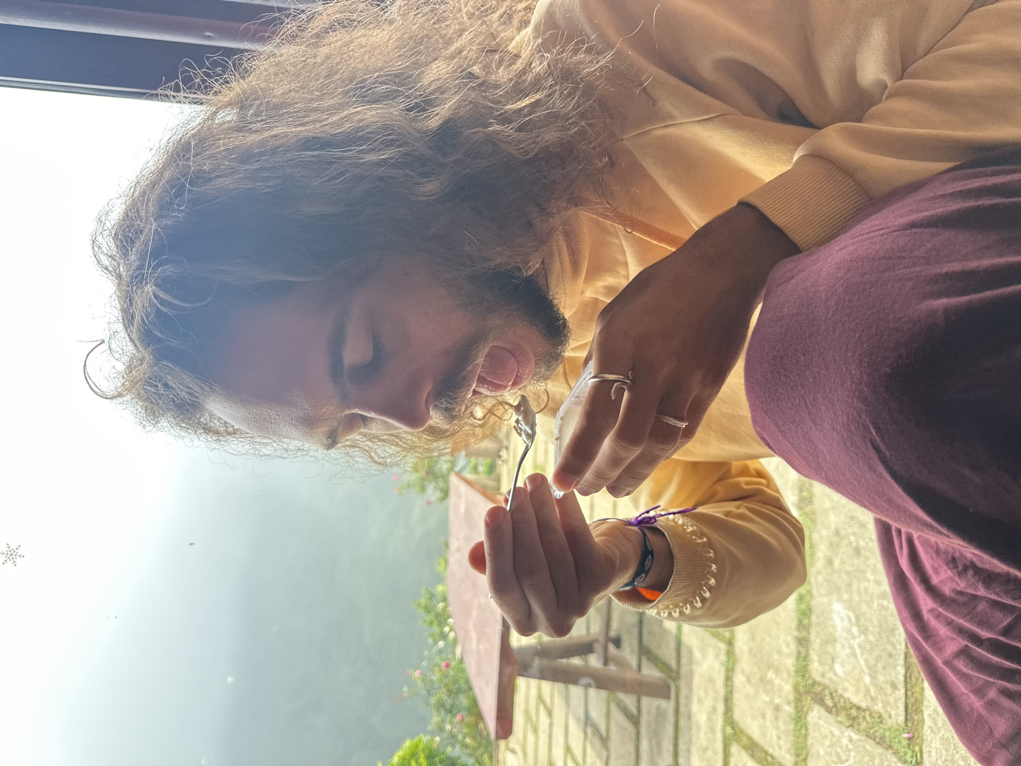 A person with long, curly brown hair and a beard is seated, holding a spoon with food towards their open mouth, preparing to eat. They are wearing a light yellow hooded sweatshirt and dark purple pants. The person is also wearing two rings on their left hand and a bracelet on their left wrist. In the background, through a window, a hazy landscape with green hills and a red table with two legs is visible. The foreground shows a stone patio. The overall scene appears to be an outdoor cafe or restaurant setting in Ta Xua, Vietnam, given the mountainous backdrop and the style of the furniture.
