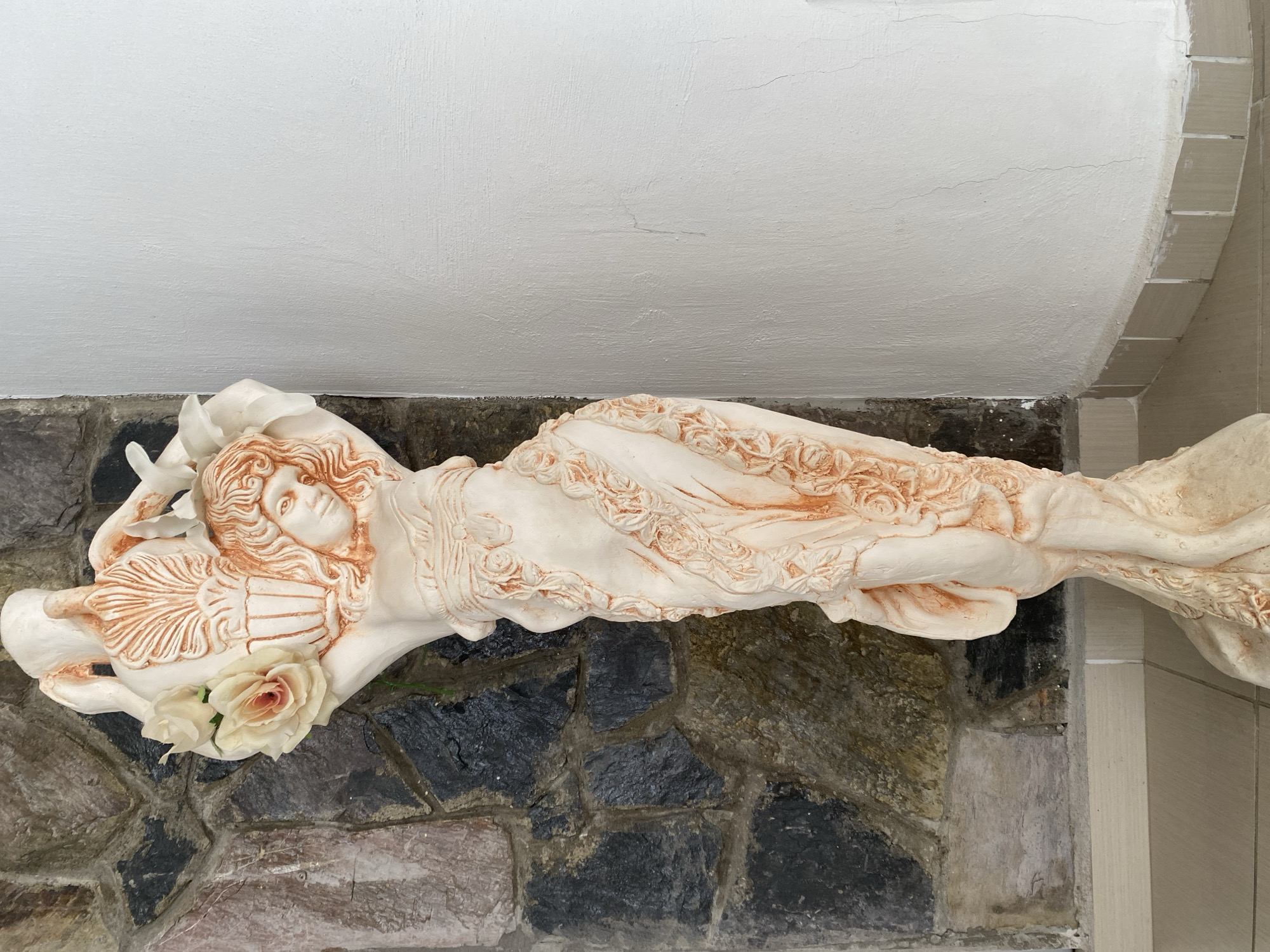 A sculpted figure of a reclining woman adorns an exterior wall in Alanya, Turkey. The sculpture, predominantly white with reddish-brown accents highlighting the details, is placed against a wall constructed from irregular, dark stones. The woman reclines on her left side, her head resting on a pillow. Decorative roses in pale colors rest near her body. The background features a curved white wall above the stone wall. The scene appears to be an architectural detail on a building exterior.