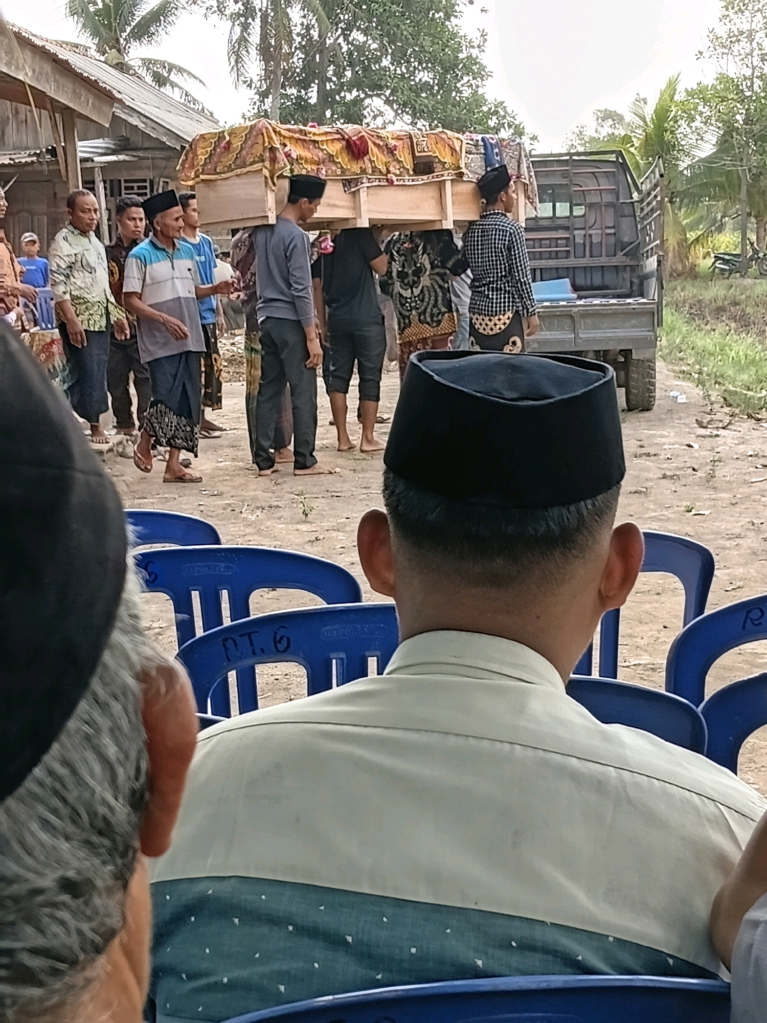 In Margo Rukun, Indonesia, a group of men are carrying a coffin draped with fabric. They are walking on a dirt path next to a small truck. A few people are watching the procession from the side, sitting on blue chairs. The scene appears to be taking place outdoors, with buildings and trees in the background.