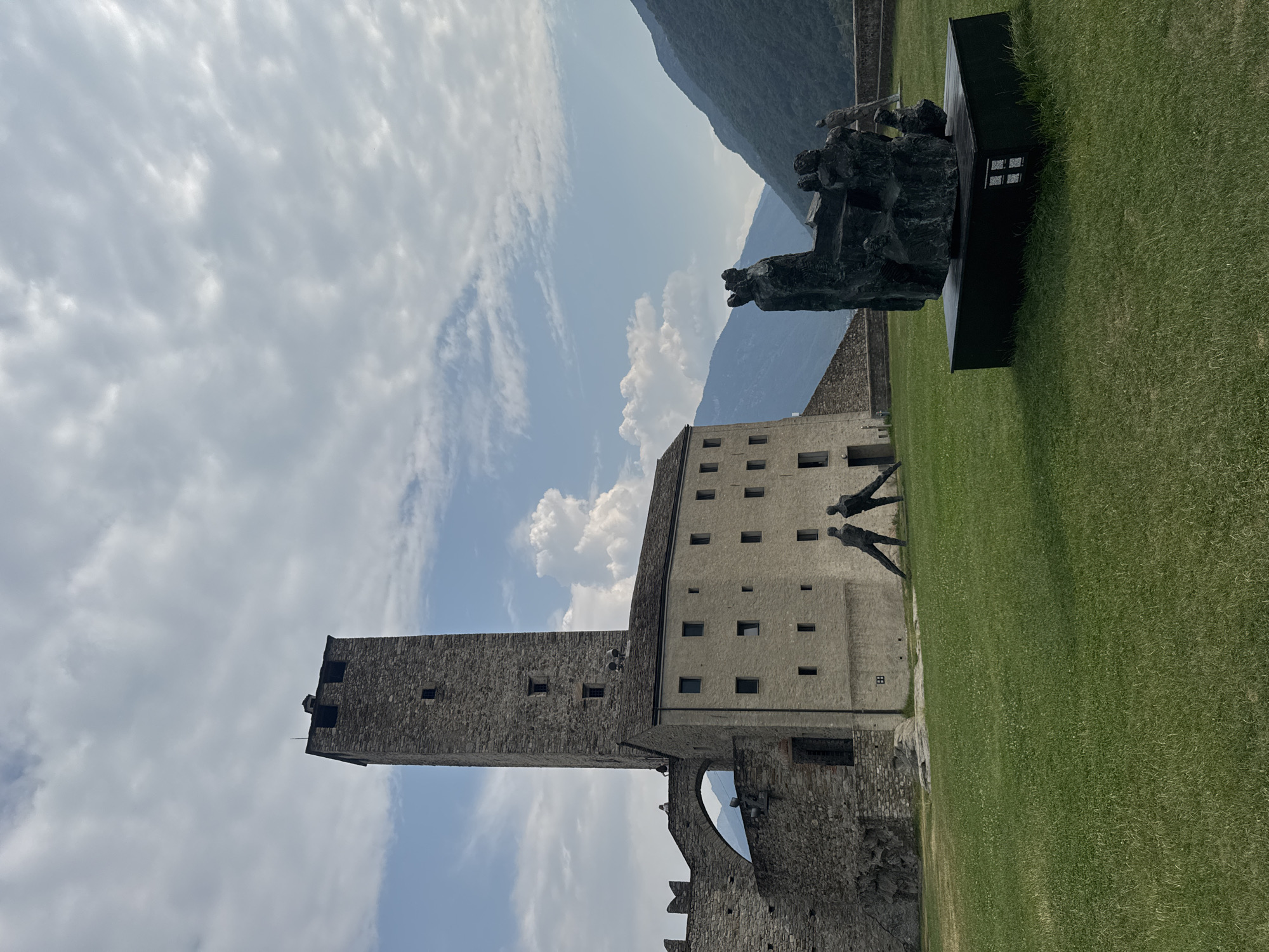The image shows the Castel Grande, a medieval castle complex in Bellinzona, Switzerland, on a partly cloudy day. The main structure is a tall, stone tower with narrow windows, attached to a long, beige building with multiple square windows. To the left, a section of the castle wall with battlements and an arched walkway is visible. In the foreground, a green lawn is dotted with a dark bronze sculpture of several figures huddled together, mounted on a black pedestal. Two smaller, dark bronze sculptures of leaning human figures are positioned on the lawn in front of the beige building. In the background, lush green mountains rise under a sky with scattered white clouds.