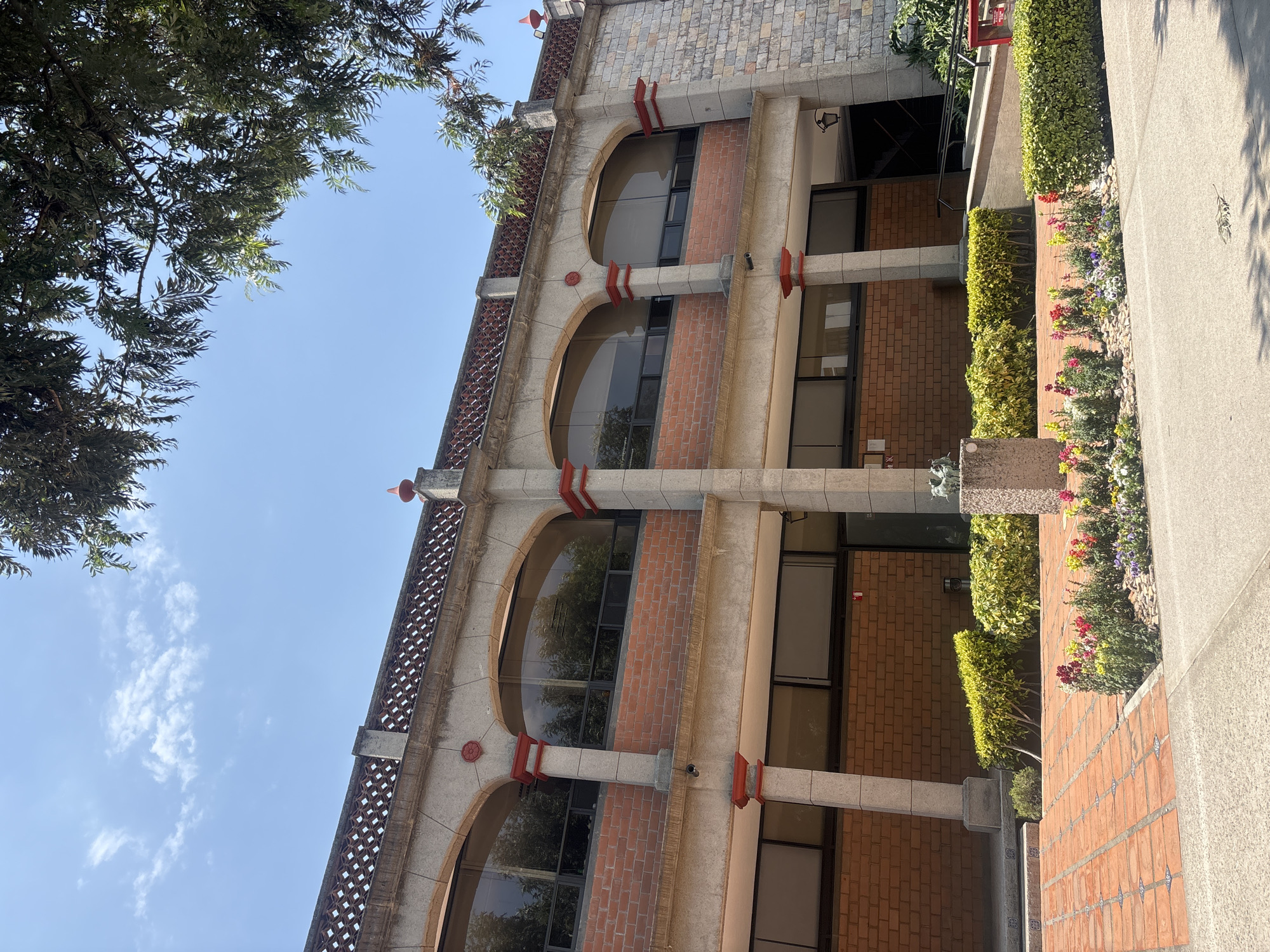 The image presents a bright, sunlit daytime view of a distinctive building and its surrounding landscape in San Andrés Cholula, Mexico. The multi-story building exhibits an architectural style, possibly Spanish Colonial Revival, characterized by a blend of light-colored stone or concrete and reddish-brown brickwork. Its facade features prominent arched windows on at least two visible floors, supported by sturdy concrete columns and adorned with decorative red-painted accents, including brackets and circular motifs. An intricate, diamond-patterned balustrade or railing system is visible along the upper edge of the building.
In the foreground, a manicured garden area runs parallel to the building. This includes a light-colored paved path, a section of reddish brick paving with small blue and white decorative tiles, and lush green hedges. A vibrant flower bed, rich with colorful blooms in reds, pinks, purples, yellows, and whites, adds a lively touch. A small, decorative sculpture, possibly a gargoyle, sits atop a concrete pillar within the garden. An entrance with dark glass doors and steps is visible towards the lower right of the building.
Overhead, a clear, brilliant blue sky, speckled with a few wispy white clouds, is partially framed by the dark green foliage of trees with slender leaves. The strong sunlight suggests a sunny day, likely mid-morning to late afternoon, evidenced by the distinct shadows cast. No people are present in the scene, conveying a sense of tranquility. There is no visible text in the image.