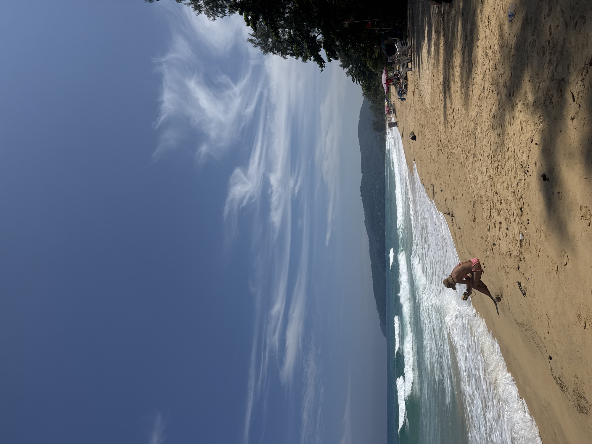 A long shot shows a sandy beach with turquoise water and breaking waves under a bright blue sky with wispy clouds. In the foreground, a woman sits on the sand facing the ocean, wearing a straw hat and a pink bikini. She is looking down at an object in her lap, possibly a book or a phone, as she holds it with both hands. The beach slopes gently upwards from the water's edge, with footprints visible in the sand. Further down the beach, there are several beach umbrellas and lounge chairs set up, with a few people scattered around. To the right, a dense line of trees borders the beach. In the distance, lush green hills form a backdrop to the scene. The overall atmosphere is one of tranquility and leisure, typical of a beach resort in Karon, Thailand.