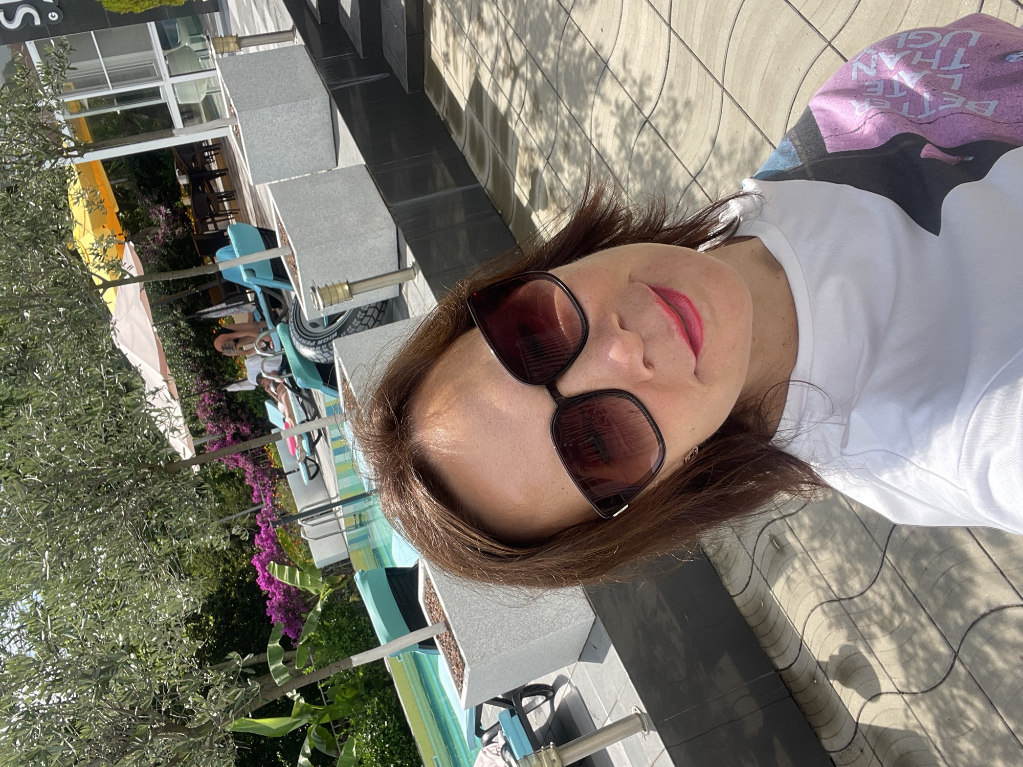 In this selfie, a woman with brown hair and sunglasses smiles while posing in an outdoor area with a swimming pool. She is wearing a white t-shirt with a graphic print on the sleeve. The background shows lounge chairs, trees, flowering bushes, and a building. The paving stones on the ground are patterned with shadows cast by trees. The scene appears to be a resort or hotel area, possibly in a warm climate given the presence of a pool and outdoor seating. The location is identified as Abaata, Abkhazia.