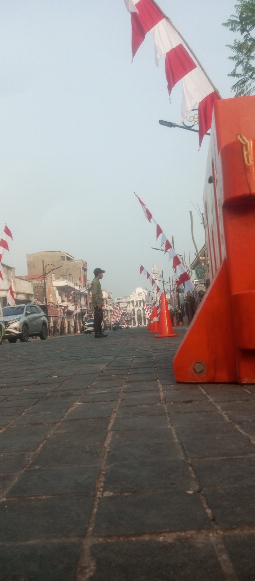 The image depicts a street scene in Kediri City, Indonesia, decorated with numerous red and white Indonesian flags. The flags are strung across the street, with some appearing tattered. In the foreground, a person wearing a dark cap and a light-colored shirt stands facing right. The street is paved with dark cobblestones, and a bright orange traffic barrier is prominently positioned on the right side of the image. Several orange traffic cones are lined up along the street. In the background, buildings with white facades and some trees are visible. Cars are present on the street, including a silver SUV on the left. The sky is overcast, suggesting it might be early morning or late afternoon. The overall impression is one of a public space prepared for or decorated for a national event.