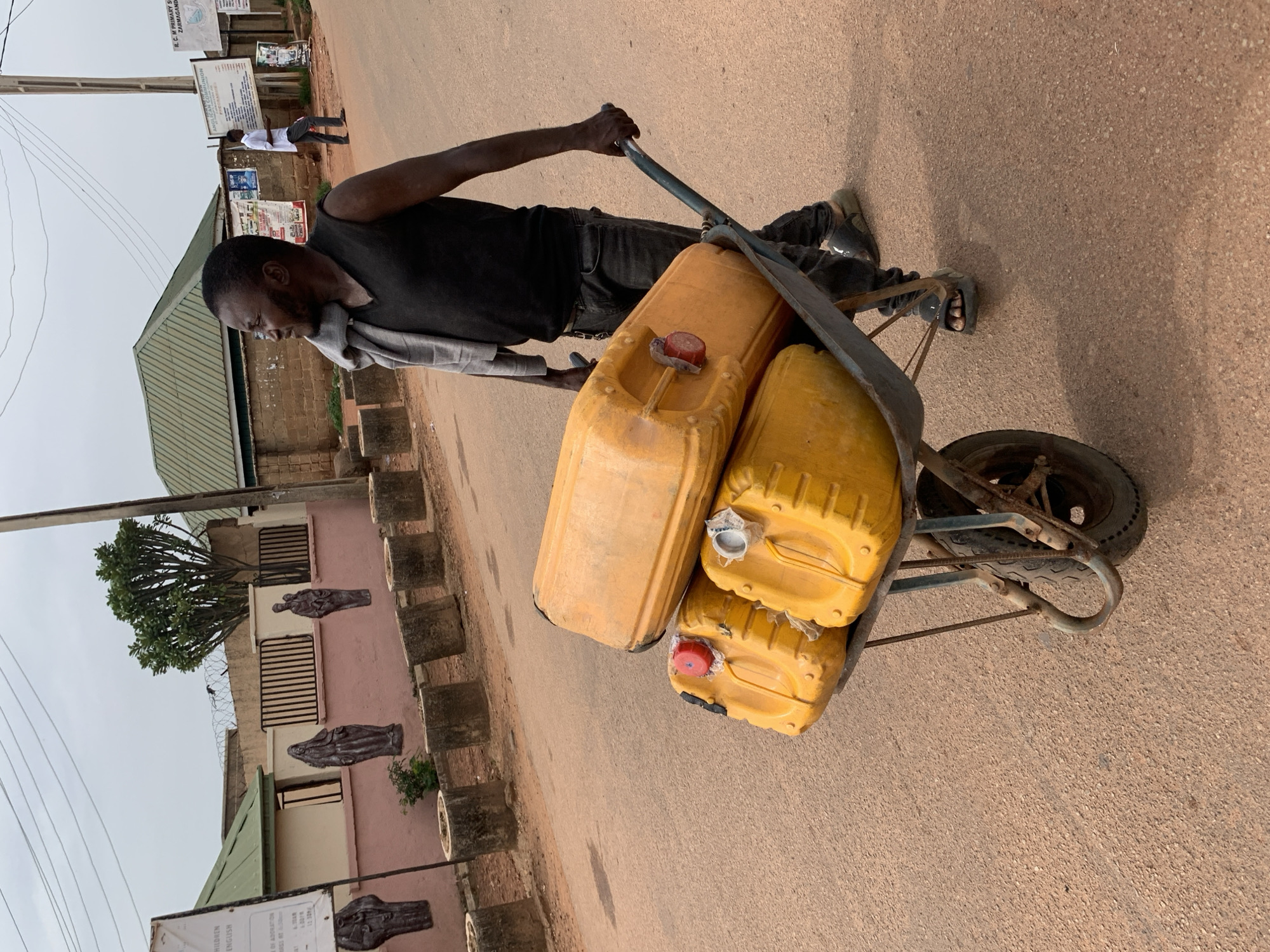 An African man pushes a wheelbarrow filled with four yellow jerrycans down a paved street in Jos, Nigeria. The man is wearing a black sleeveless shirt, dark pants, and sandals. He has a gray cloth draped over his left shoulder. The jerrycans are stacked, with one large one on top and three smaller ones below. The wheelbarrow has a single wheel and metal handles. In the background, there is a building with a pink wall and green roof, a fence, and trees. A utility pole stands to the right of the frame. The sky is overcast.
