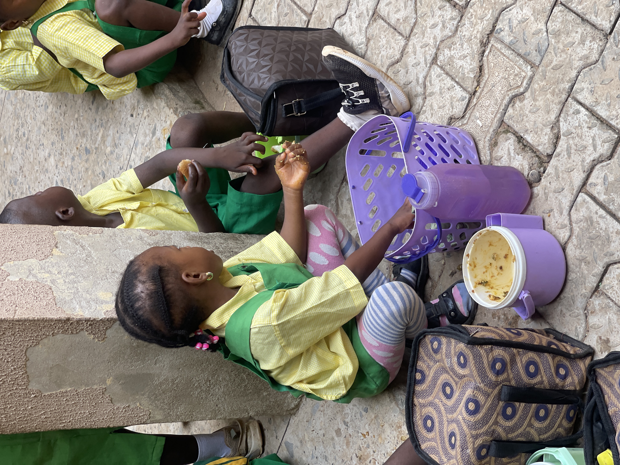 In the city of Akwanga, Nigeria, several children in school uniforms are seated on a paved area, appearing to be during a meal break. A young girl in the foreground, wearing a yellow plaid shirt with a green vest and striped leggings, is crouched down. She has her hair in braids with pink beads and is holding food in her hands, with some on her fingers. Beside her, a purple plastic basket and a purple water bottle are visible. To her left, a patterned lunch bag rests on the ground. In the background, two other children are also seated, one of whom is holding a piece of bread. The environment consists of a textured wall and a paved ground.