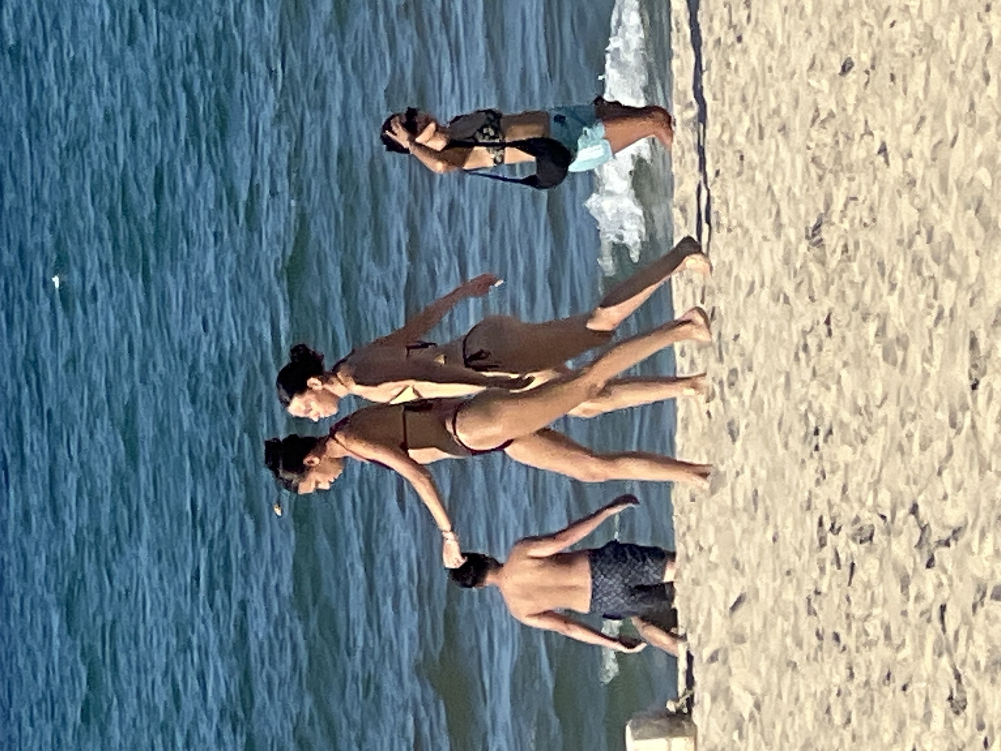 This image captures a sunny day at My Khe Beach in Da Nang, Vietnam. The scene features four individuals by the water's edge. In the foreground, two women are walking on the sand. The woman on the left is wearing a dark bikini and has her hair pulled up. The woman on the right is also in a bikini, with her hair in a bun. Behind them, a man in patterned swim shorts stands in shallow water, with his back to the camera. Further to the right, another woman in a bikini top and blue shorts stands near the shoreline, holding her hand to her forehead and wearing sunglasses. The ocean, with its blue-green water and gentle waves, forms the backdrop. The sandy beach is textured with footprints.