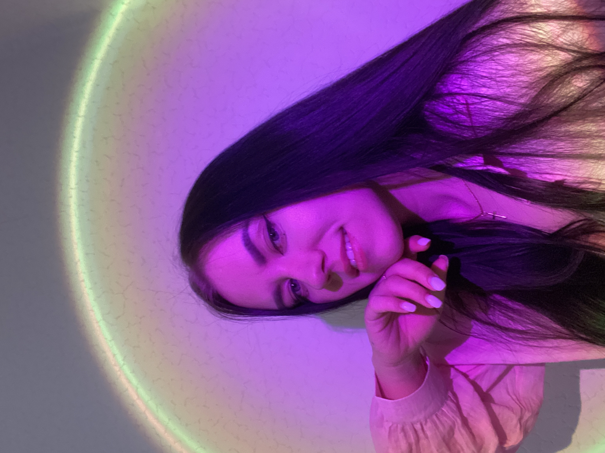 The image shows a young woman smiling and looking directly at the camera, her head tilted to the side, possibly indicating she is lying down or positioned at an angle. She has long, dark hair that cascades around her. She appears to be wearing a light-colored, possibly peach or light pink, top with ruffled details on the sleeves. Her fingernails are painted a pale shade. A delicate necklace with a small cross pendant is visible around her neck.
The background is a wall illuminated by vibrant artificial lighting, creating a distinct atmosphere. A prominent circular or arched light pattern, transitioning from bright green/yellow to purple/pink, is projected onto the wall, likely from a sunset lamp or similar device. The rest of the wall and her face are bathed in a strong purple and pink glow. The wall itself has a subtle, textured appearance with small, irregular speckles. The overall mood conveyed is relaxed and engaging. There is no visible text in the image. The location context for this scene is Chaikovsky, Russia.