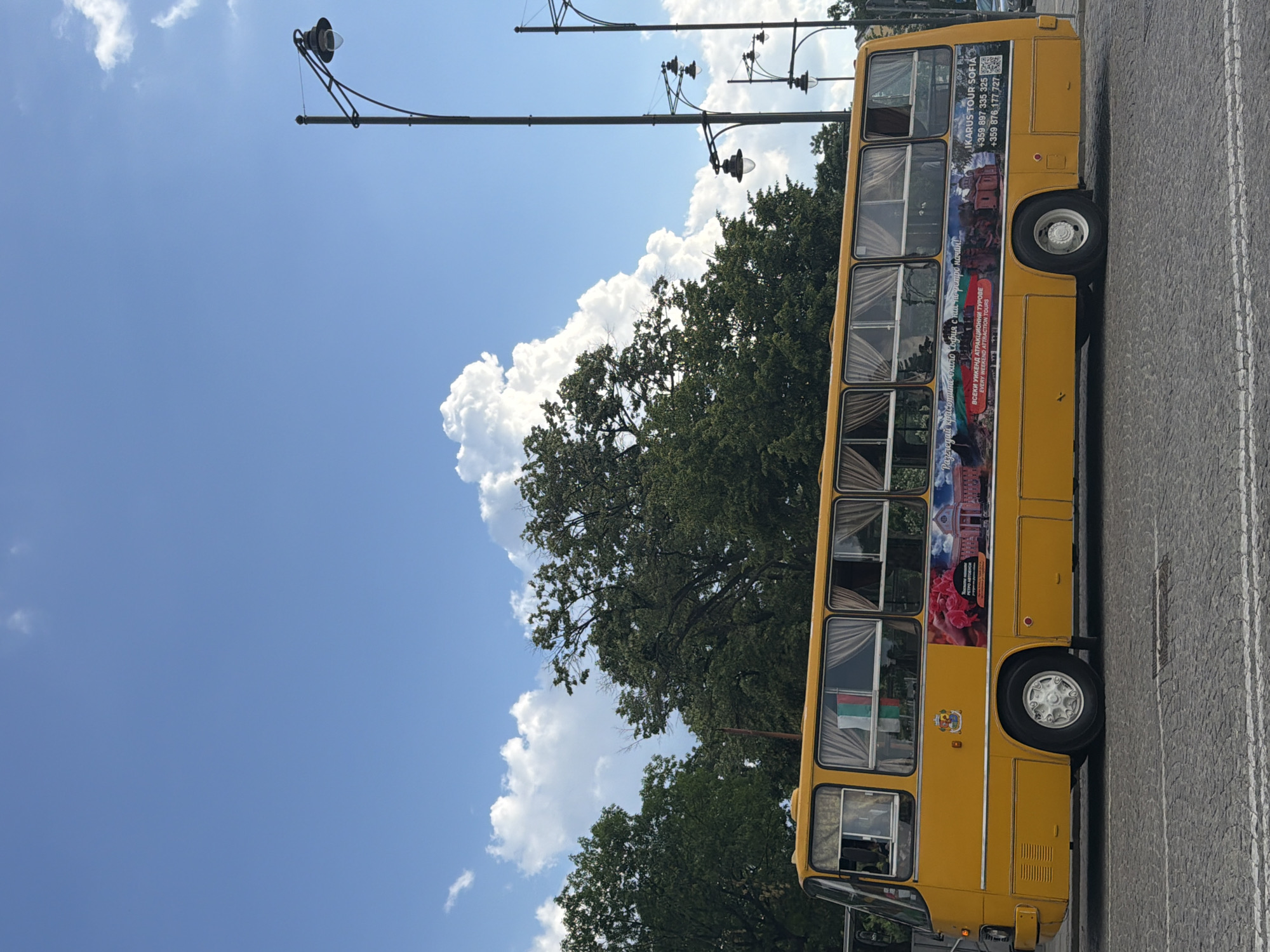 A bright yellow retro-style Ikarus bus is parked on a cobblestone street in Sofia, Bulgaria, viewed from its left side. The bus features multiple rectangular windows, each fitted with light-colored curtains drawn inward. A small Bulgarian flag (white, green, red horizontal stripes) is visible through one of the front-left windows. Below the front passenger window, a crest or emblem is affixed. The side of the bus displays large advertisements with images including red roses, architectural landmarks, and various Bulgarian landscapes. Text on these advertisements, in both Bulgarian and English, promotes "IKARUS TOUR SOFIA," offering "retro" and "attraction tours" with contact phone numbers (+359 897 335 325, +359 876 177 727) and a QR code. The bus has white hubcaps on its visible wheels. The background features a dense canopy of lush green deciduous trees. The upper half of the image is dominated by a clear blue sky with scattered, fluffy white cumulus clouds, including a particularly large formation on the left. Multiple black, ornate streetlights with globe lamps are positioned vertically on the right side of the frame. No people are visible in the scene.