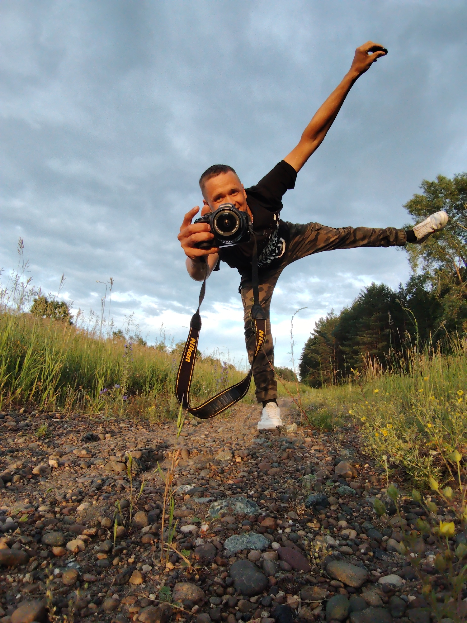A man is captured mid-action, holding a Nikon camera in his right hand and taking a photo of the viewer. He is wearing a black t-shirt, camouflage pants, and white sneakers. His left arm is raised in the air, and his right leg is extended horizontally behind him, showcasing a dynamic pose. The scene appears to be outdoors, with a gravel path in the foreground and tall grass and wildflowers on either side. In the background, a dense line of trees is visible against a cloudy sky. The lighting suggests it is late afternoon or early evening. The man's expression seems joyful and energetic. The camera strap, with "Nikon" clearly visible, dangles around his neck. The overall mood is playful and spontaneous.