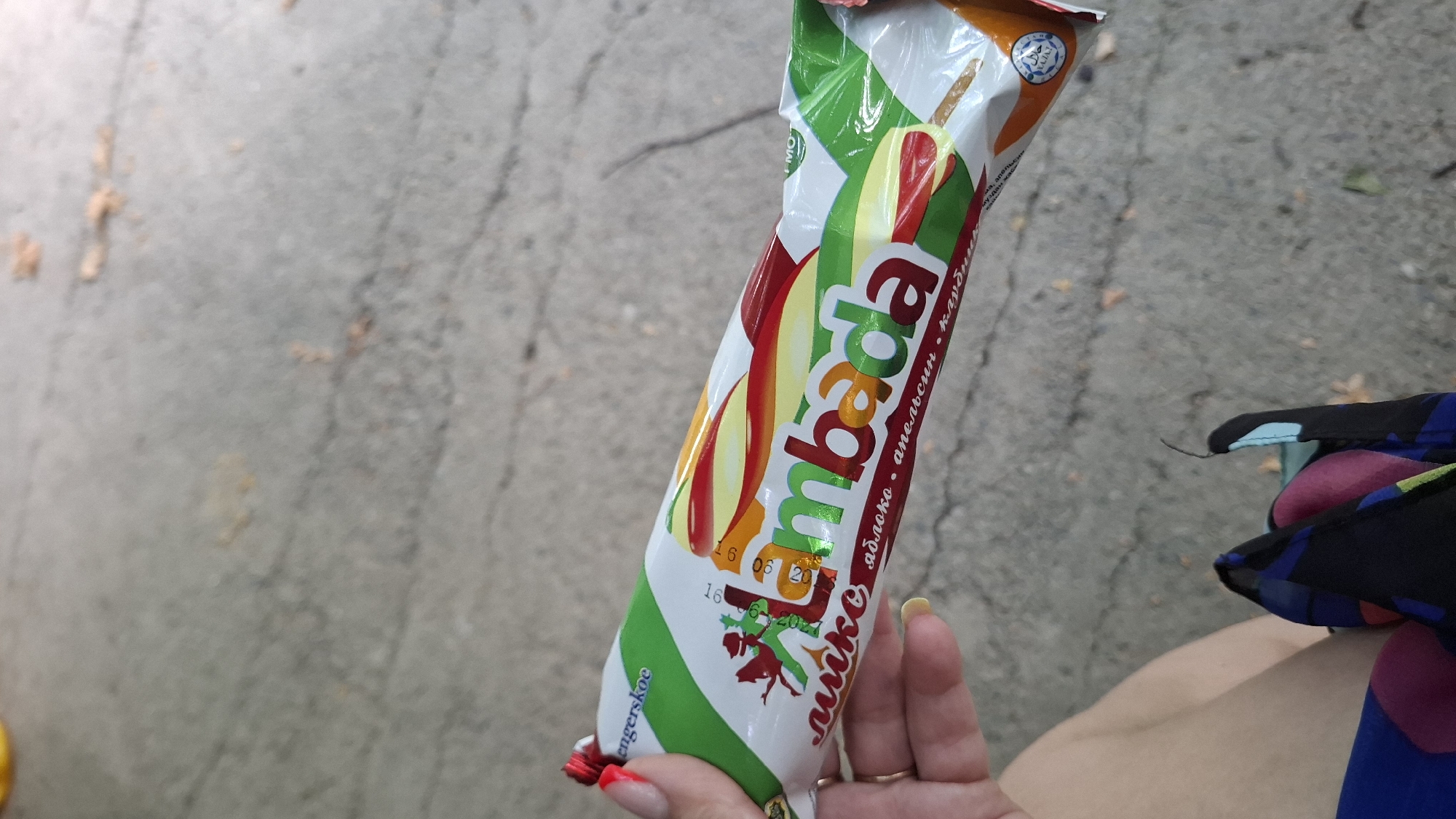 A person's hand with bright red nail polish is holding a wrapped ice cream bar. The ice cream bar has a white wrapper with green and colorful fruit imagery, and text that appears to be in Cyrillic script. The brand name "Lambada" is prominently displayed in large, multicolored letters on the wrapper. Below the brand name, there are words that likely describe the flavors. The background consists of a textured gray surface, resembling pavement or concrete, with some scattered organic debris. A small portion of the person's leg and a colorful scarf or fabric are visible in the lower part of the image.