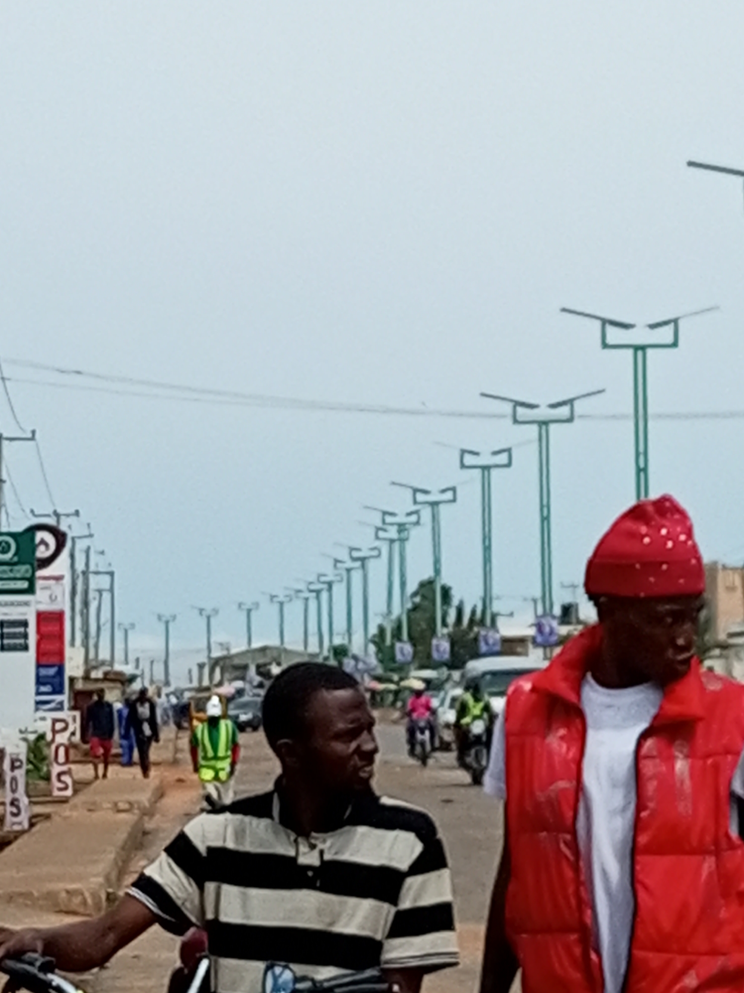 The image depicts a street scene in Garko, Nigeria, characterized by a row of modern streetlights lining both sides of the road. In the foreground, two men are prominent. The man on the right wears a red puffer vest over a white t-shirt and a red head covering adorned with white beads. The man on the left, partially obscured by the motorbike he is on, wears a black and white striped t-shirt. In the background, several other individuals are visible walking and riding motorcycles. One person in the middle ground wears a bright green vest, suggesting they might be a worker or security personnel. Vehicles, including cars and motorcycles, are present on the road. The environment appears to be an urban or semi-urban area with buildings and shops lining the street. The sky is overcast, suggesting a cloudy day. Signs, including one with "POS" visible, indicate commercial activity. The overall impression is of a bustling street in a Nigerian town.