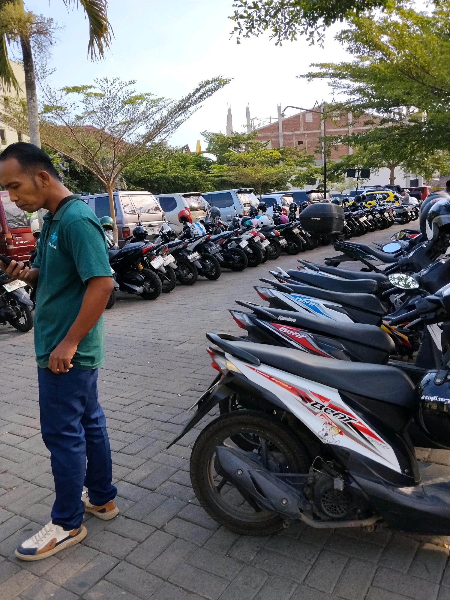 A man in a green polo shirt and blue pants stands on a paved walkway next to a row of parked motorcycles. He is looking down at his phone, which he holds in his left hand. The man has short, dark hair and a mustache. In the background, more motorcycles are parked alongside several vehicles, including vans and cars. A building under construction is visible in the distance, as are trees and a clear sky. The scene appears to be in a public parking area in Demangan, Indonesia.
