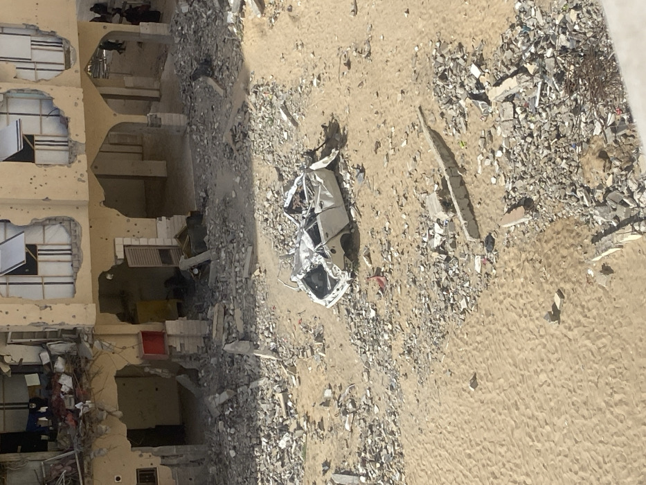 An overhead view captures a scene of urban destruction in Khan Yunis, Palestinian Territory. The foreground consists of sandy ground heavily interspersed with concrete rubble, fragmented building materials, and scattered debris. A severely damaged, gutted white sedan chassis, missing its wheels, roof, and interior components, rests centrally in this debris field. In the background, a multi-story building, likely a former residential or commercial structure, exhibits extensive damage. Its light-colored facade is pockmarked with numerous circular impact holes, suggesting projectile strikes. Upper-floor windows are either shattered or crudely boarded with makeshift white coverings and wooden frames. Laundry, including blue and white garments, hangs from a clothesline on an exposed upper balcony. The ground floor features a series of arched openings and exposed rooms, filled with more debris. A red rectangular object is attached to a ground-floor pillar, and a yellow object is visible within one of the open ground-floor spaces. In the far background, several dark-clothed figures, appearing to be adults, are observed walking near the damaged building. The scene is illuminated by natural daylight.