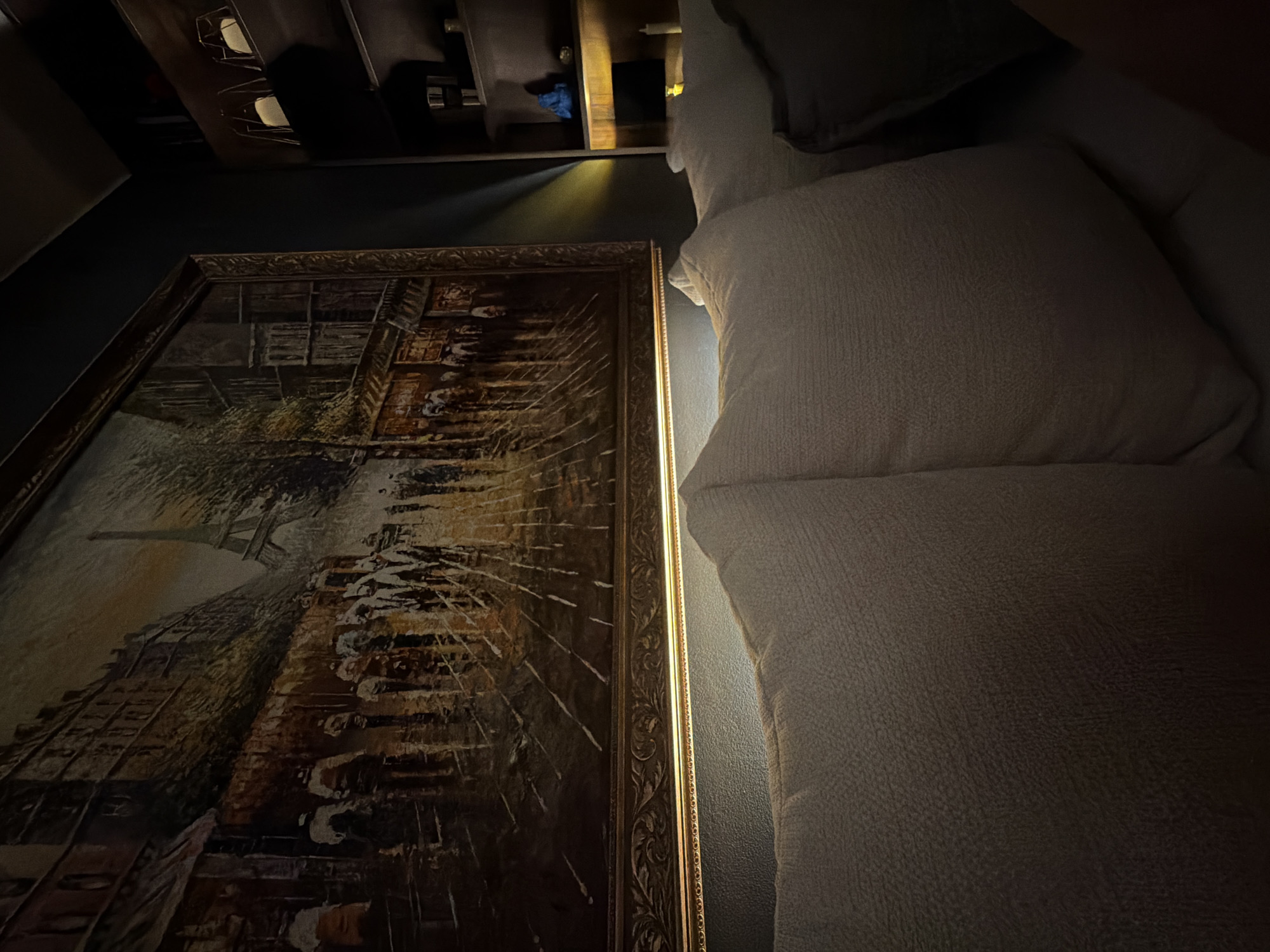 The image displays a dimly lit interior scene featuring a large painting of a Parisian street with the Eiffel Tower in the background. The painting is prominently displayed on a wall above several plush, light-colored sofa cushions. To the right of the painting, a dark bookshelf is visible, containing various objects including decorative lamps, a blue figurine, and other items that are difficult to discern due to the low lighting. A soft, yellow light emanates from a spot on the wall below the painting, illuminating the edge of the sofa. The overall ambiance suggests a cozy, indoor setting, likely a living room or lounge area. There is no indication of the city of Plovdiv, Bulgaria, in this image.