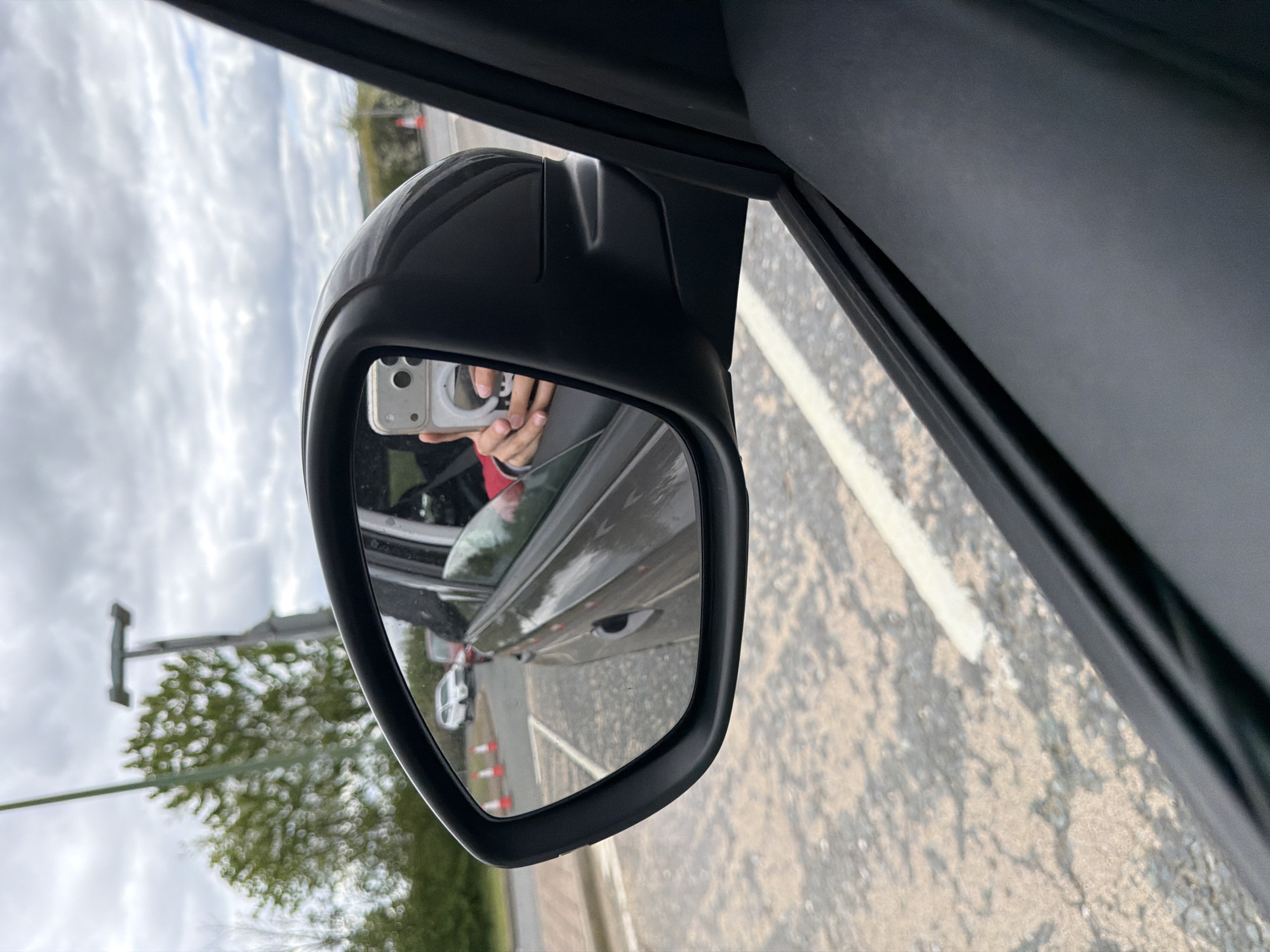 The image shows a side view mirror of a vehicle, reflecting the driver taking a photograph of the mirror with their own smartphone. Reflected in the mirror is the side of the vehicle to which it is attached, a line of traffic cones, a parked white car, an electrical pole and a tree. The road surface is visible outside of the vehicle, marked with a white line. The location is identified as Pity Me, United Kingdom.