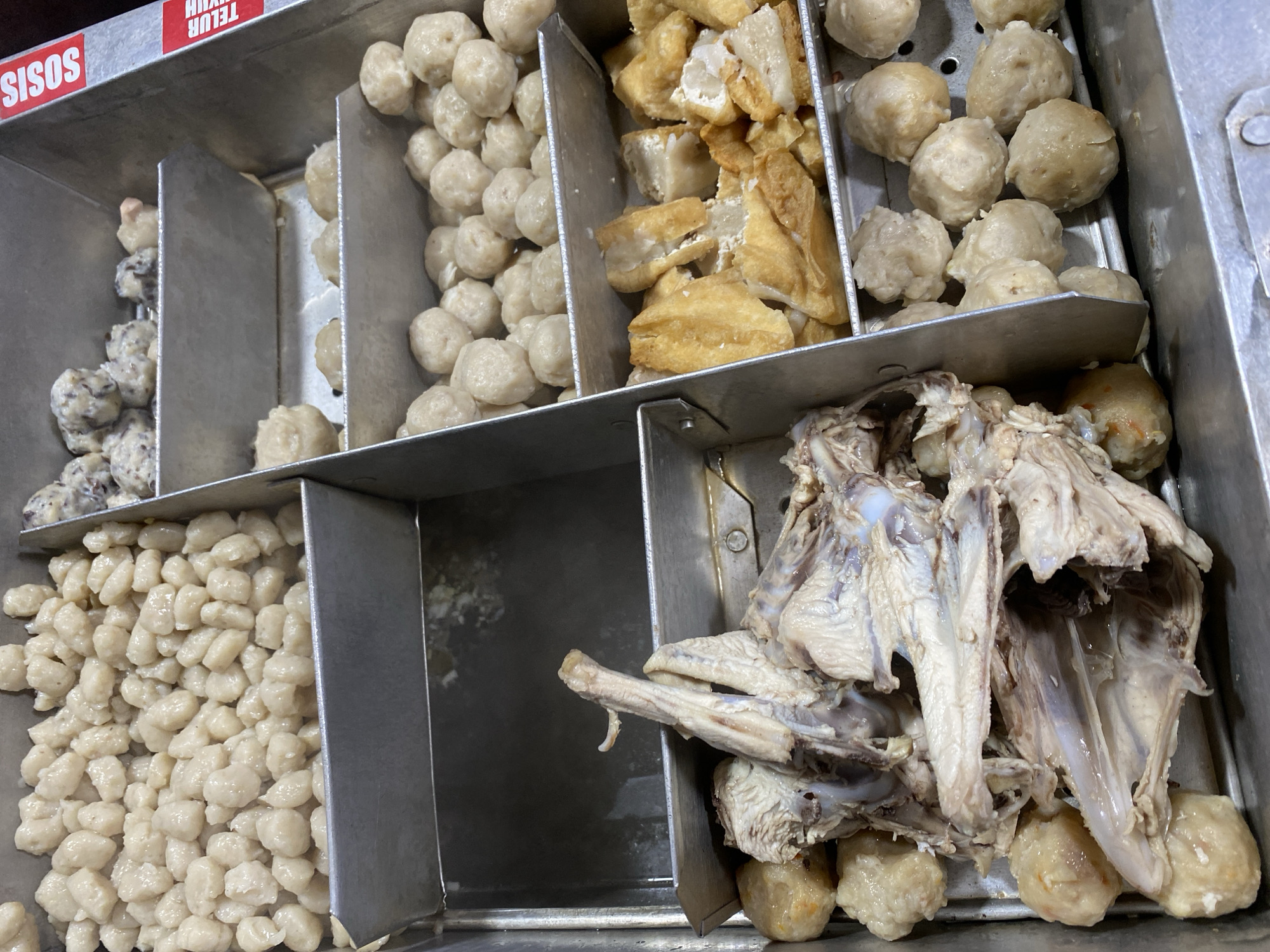 A metal bin is filled with various food items. In the top left section, small, pale, irregularly shaped balls of food are stacked. To their right, in the top right section, are more balls of food, some appearing slightly darker and mixed with what looks like finely chopped ingredients. The middle section contains larger, round, pale balls of food, some of which have slight browning. Below these, on the left, is a pile of cooked chicken carcass with some meat still attached, interspersed with pale, round food balls. In the bottom right section, there are golden-brown, fried pieces of tofu. The bin is made of stainless steel and has dividers. A red label with white text, reading "SISOS" and "TELUR AYAM", is visible on the right side of the bin. The background is dark. The image appears to have been taken at eye level, looking down into the bin. The scene is likely a food stall or market, possibly in Kasihan, Indonesia, given the labeling.