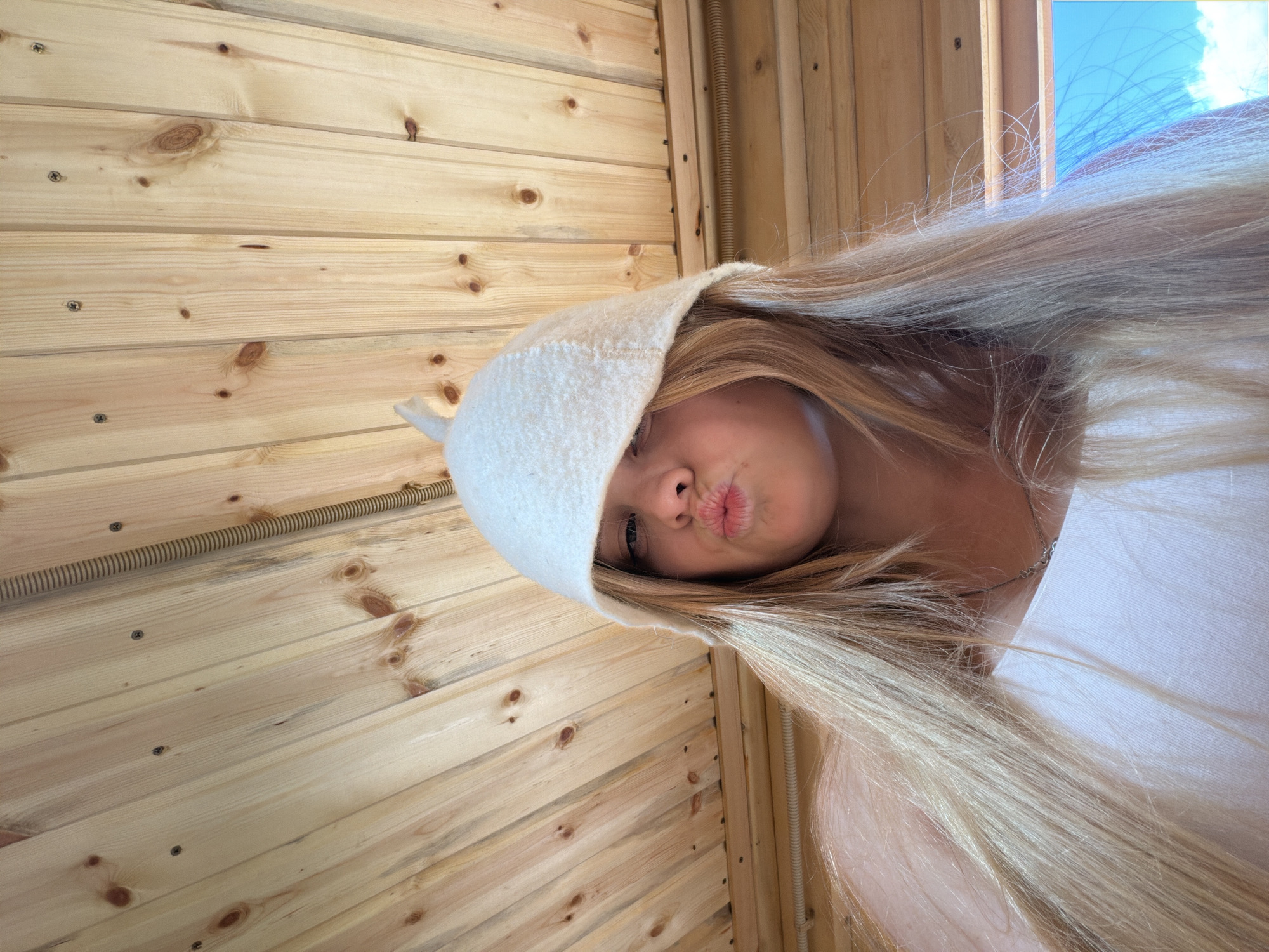 This image captures a close-up, angled selfie of a person with long, flowing blonde hair, partially covered by a white felt sauna hat. The person is making a playful "pouty" or "kissing" face towards the camera, with their eyes, nose, and mouth visible beneath the hat's brim. A faint silver chain is visible around their neck, and they appear to be wearing a light-colored top.
The setting is characterized by light-colored wooden paneling, likely pine, forming the background. The wooden planks display natural knots and are secured with visible screws. A corrugated or coiled tube, possibly an electrical conduit or part of a heating system, runs vertically along one section of the wooden wall. The presence of the distinctive white felt hat strongly indicates the scene is set in a sauna, potentially in Vozhoiskii, Russia.
Bright daylight illuminates the scene, with a glimpse of a blue sky and white clouds visible through an opening or window in the upper right, suggesting it is daytime. The overall mood conveyed is casual and lighthearted. No discernible text is visible within the image.