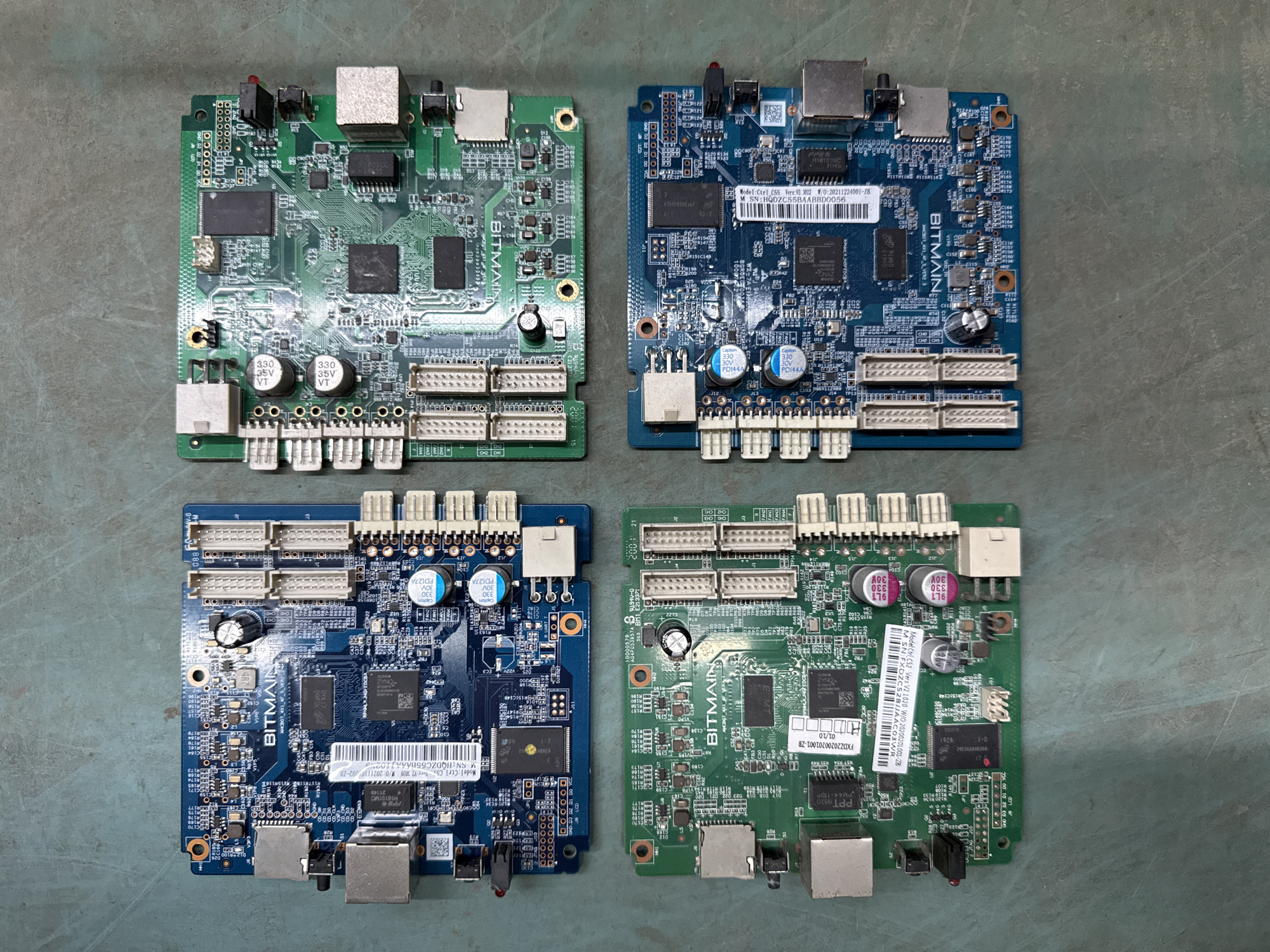 Four electronic circuit boards are laid out on a textured, grey surface. Each circuit board is rectangular and features various electronic components, connectors, and printed text.
The top-left board is blue and has "BITMAIN" printed in white lettering. It includes an RJ45 connector, several smaller connectors, and numerous chips and resistors. A white label with a barcode and text is visible.
The top-right board is green and also has "BITMAIN" printed on it. It features two large black chips, smaller components, and several connectors.
The bottom-left board is green and has "BITMAIN" printed on it. Similar to the top-left board, it has an RJ45 connector, smaller connectors, and a white label with a barcode and text. Two capacitors are prominently visible near the top edge of the board.
The bottom-right board is blue and has "BITMAIN" printed on it. It features two large black chips, smaller components, and several connectors, including an RJ45 connector. A white label with a barcode and text is visible.
The grey surface appears to be a floor or a workbench. The lighting suggests an indoor environment. No people, animals, or any other objects are present in the image. The image is taken from a top-down perspective.
The context of Indiantown, United States of America, is not discernible from the visual information in the image.