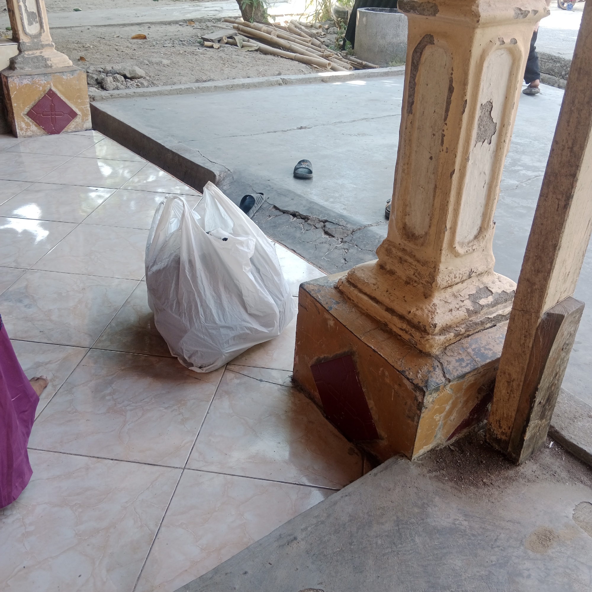 In the city of Tegowanu Kulon, Indonesia, an image captures a tiled outdoor area adjacent to a concrete floor. A large white plastic bag, likely containing some contents, sits on the tiled surface. To the right of the bag, a weathered, pillar-like structure with chipped paint stands. In the background, a pile of bamboo poles is visible on a dirt patch, and further beyond, there is a cement pipe. A pair of dark-colored flip-flops is on the cracked concrete floor. A portion of a person wearing a purple garment is visible in the lower left corner of the image.
