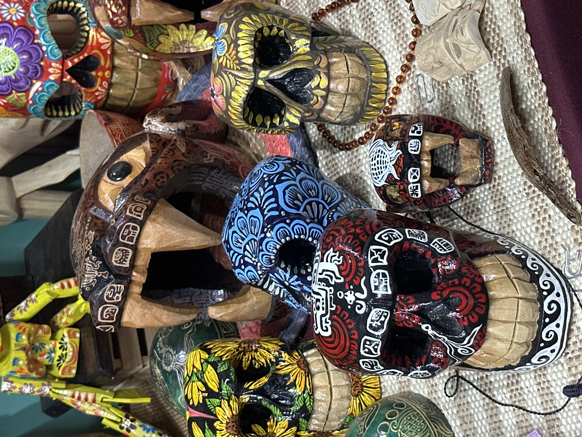 The image presents a collection of vibrantly decorated wooden skulls and animal masks, typical of Mexican folk art. The skulls are adorned with intricate designs featuring flowers, geometric patterns, and indigenous motifs in a variety of colors including red, blue, yellow, black, and white. Some of the skulls have articulated jaws. A wooden animal mask, possibly a jaguar, is also visible with intricate carvings. The items are arranged on a textured surface, possibly a woven mat or fabric, with a few small wooden carvings, and beaded necklaces. The scene likely represents a craft market or artisan's display, with the items available for purchase. Given the style and subject matter, it's plausible that this scene could be located in a market or shop in Mexico City, Mexico, known for its rich tradition of Day of the Dead crafts and folk art.