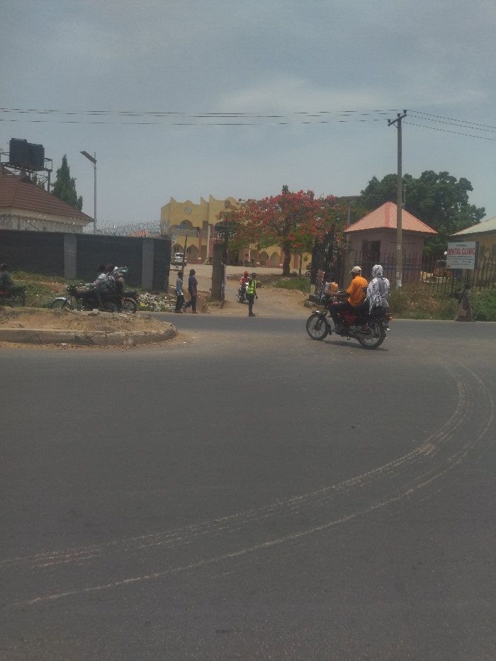 The image depicts a street corner in New Karu, Nigeria. A motorcycle with two people on it is turning onto the street from a smaller side road. The motorcycle is driving on the right side of the road. There are several other people walking on the side of the road. There is a small building with a sign for "Dental Clinic". There is a large tree with red flowers in the background. The sky is overcast. The street is paved and there are tire tracks on the pavement. There is some debris on the side of the road. The location is a residential area with houses and buildings on both sides of the street. There is a metal gate in the background, and the view is from a slight elevation, giving a perspective down onto the street. The street is relatively empty, except for the people and motorcycle.