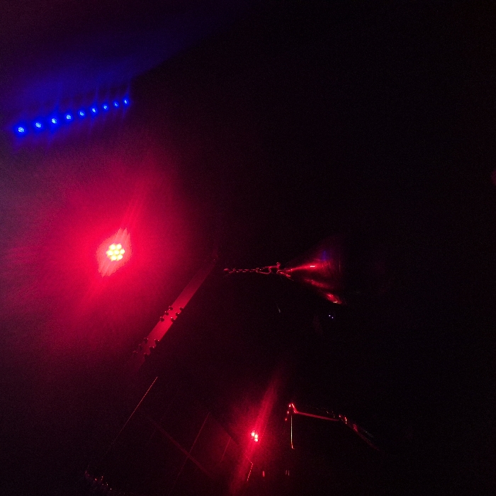 The image displays a dimly lit indoor environment characterized by artificial red and blue lighting. A dark, conical speed bag or punching bag hangs centrally from a horizontal metal structural beam via a chain suspension. The bag reflects a red light from an overhead multi-LED red fixture, which shows at least six individual emitters, positioned slightly above and to the right of the bag. Another smaller red light source illuminates equipment on the far left. A vertical strip of multiple blue LED lights is visible in the upper right quadrant. No people, actions, or interactions are present. The precise location within Strasbourg, France, is not identifiable from the visual content provided.