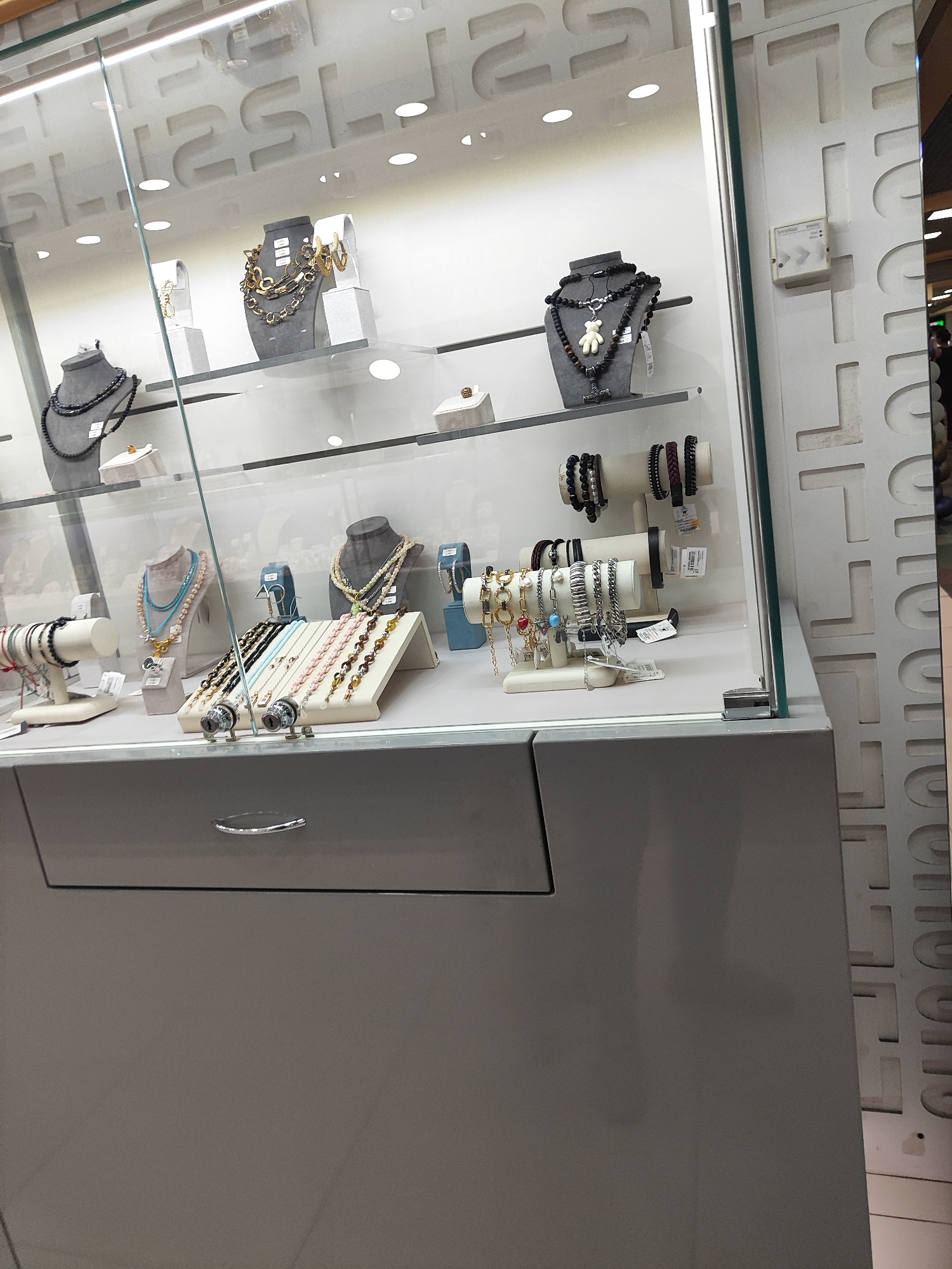 The image shows a display case filled with jewelry, likely in a retail store. The jewelry includes necklaces, bracelets, and possibly earrings. The display case has multiple shelves and is illuminated from above. The overall environment appears to be indoors, with a modern aesthetic.