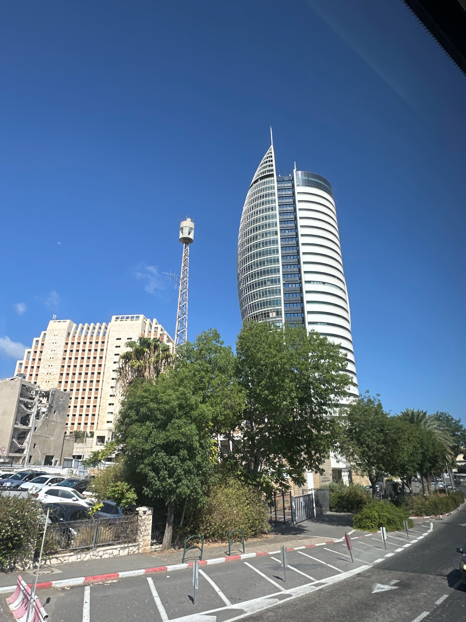 In a bright, sunny outdoor scene, a clear blue sky serves as the backdrop for a cityscape in Haifa, Israel. Dominating the right side of the frame is a tall, modern, white, and glass skyscraper with a distinctive curved facade and a pointed spire. To the left of this skyscraper is a much older, stepped building constructed of light-colored stone with rows of dark windows, appearing somewhat dilapidated with exposed concrete and scaffolding visible on its lower floors. Between these two prominent structures, a tall, slender lattice tower with a cylindrical object at its top stands against the sky, possibly a communication tower. Lush green trees and foliage are visible in the mid-ground, obscuring the bases of the buildings and the parking lot. In the foreground, a street with white lane markings and red-and-white curbs is visible, with several cars parked along the side. The overall impression is one of urban development juxtaposed with greenery and elements of older infrastructure.