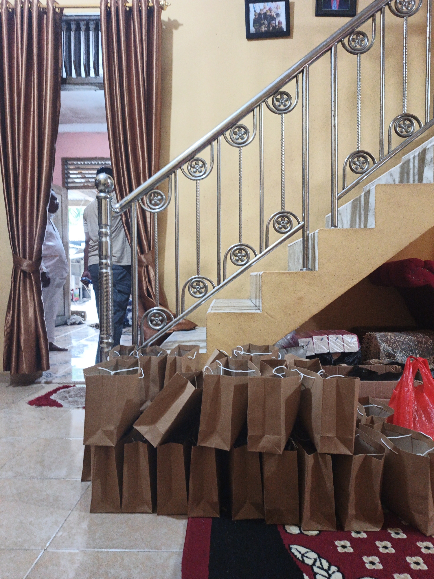 The image depicts the interior of a residence in Pematang Duku, Indonesia, featuring a staircase with a metal railing. In the foreground, numerous brown paper gift bags are stacked on the floor, some appearing to be empty. A red plastic bag is visible to the right, and beyond the staircase, several packages are stacked. Two individuals are partially visible in the background, one standing near an open doorway and another further inside the house, suggesting an indoor gathering or event. The environment appears to be a home, possibly set up for a celebration or to distribute items.