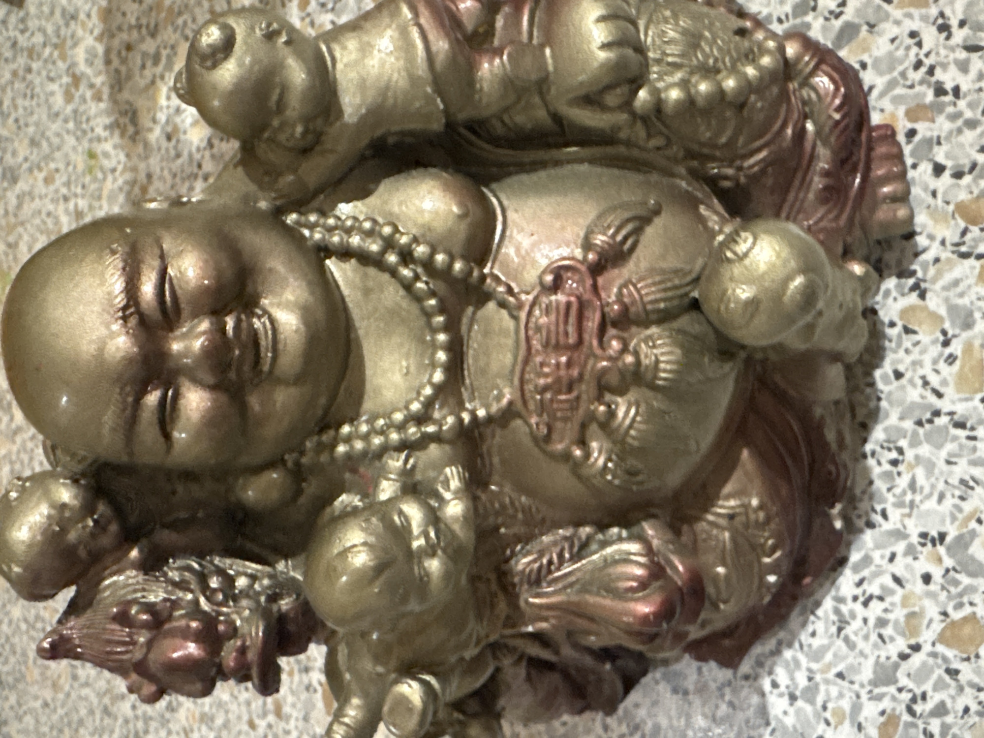 A close-up, eye-level shot captures a golden and reddish-brown figurine of a laughing Buddha, adorned with a pearl necklace. The Buddha is surrounded by several cherubic figures, with one on his left shoulder and another emerging from his left armpit. A dragon head peeks out from the upper left of the figurine. The Buddha's belly is prominent, and a decorative element hangs from his chest, featuring what appear to be Chinese characters in red. The figurine rests on a speckled, light-colored surface.