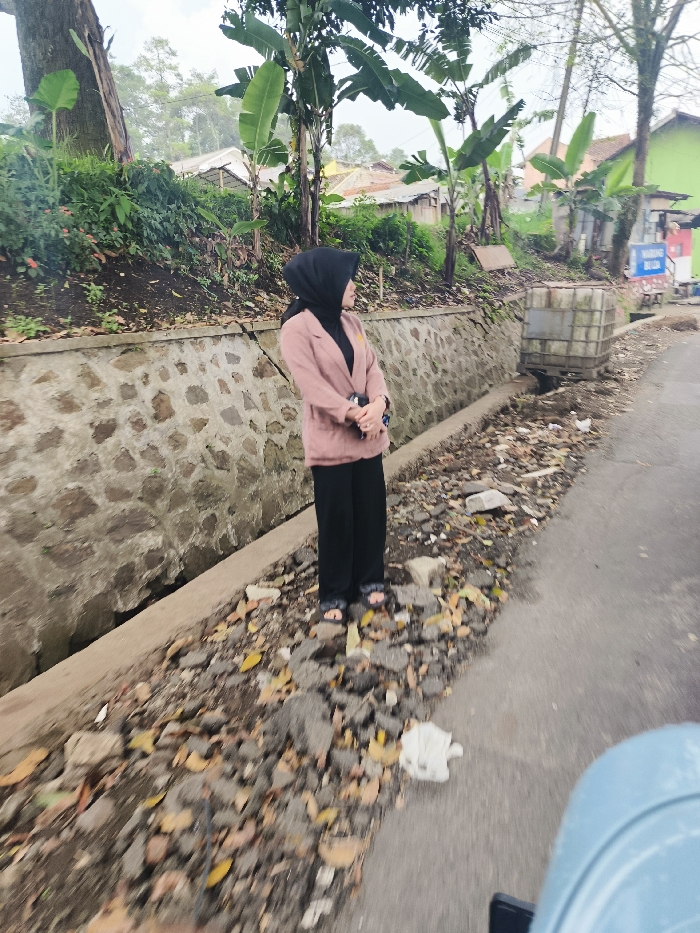 The image shows a woman wearing a black hijab and a pink blazer standing on a paved road in Lembang, Indonesia (latitude: -6.835, longitude: 107.602). The woman is facing away from the camera and is looking to the right. She is standing on a section of the road that has been partially covered in gravel. Behind her is a low, stone wall topped with a concrete curb. There are trees and houses visible behind the wall. The road is surrounded by vegetation and debris. The image is taken from a low angle, giving the impression of a perspective from a vehicle. The image appears to be taken in daylight, with a cloudy sky. There is a small, gray metal box in the background next to the stone wall. There is a light blue object in the foreground of the picture that appears to be a vehicle, possibly a car, but it is partially obscured from view.