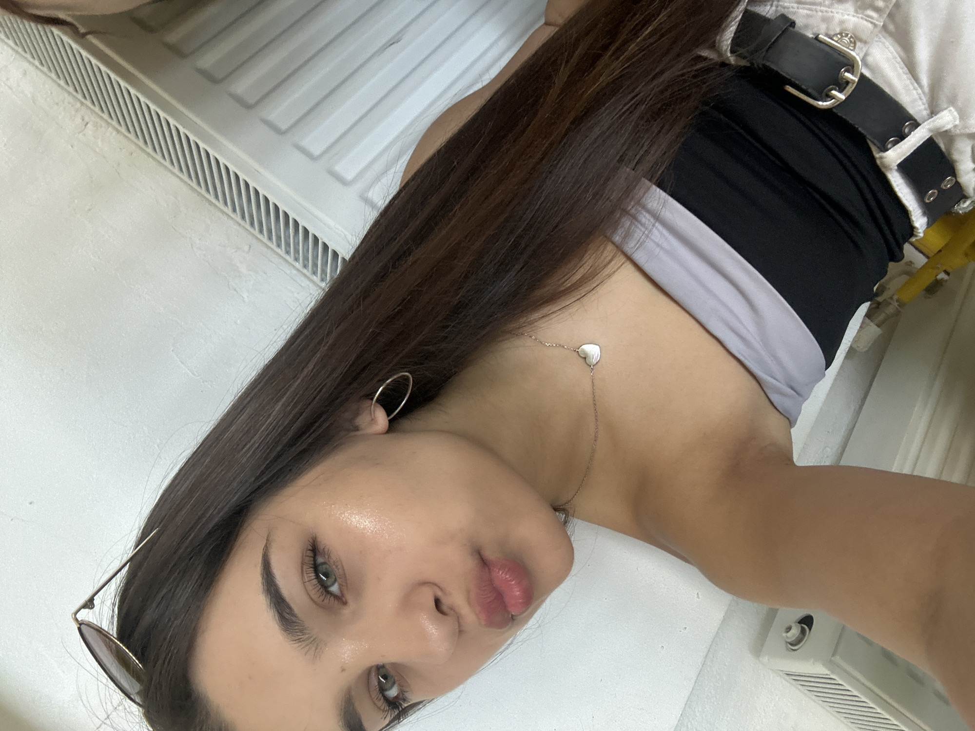 A young woman with long, dark brown hair and green eyes is taking a selfie. She is wearing a strapless top with a grey and black pattern, a silver heart necklace, and a black belt over light-colored pants. Her lips are pursed, and she is wearing sunglasses pushed up on her head. The background appears to be an indoor setting with white walls and a white radiator. The image was taken in Shumen, Bulgaria.