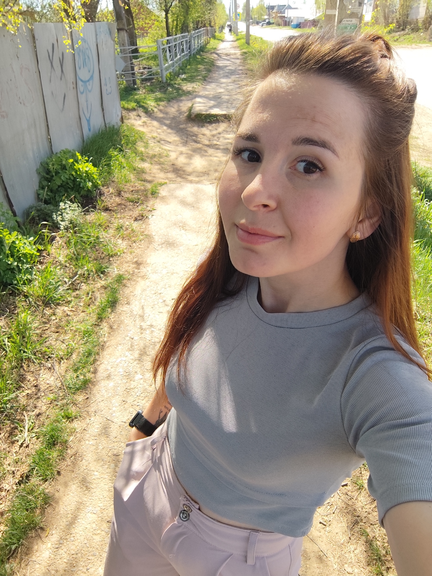 A woman is taking a selfie on a path in Фоки, Russia. The path is made of dirt and gravel, with grass and vegetation growing along the edges. A concrete wall with graffiti is visible on one side of the path, and a metal fence runs along the other side. In the background, there are trees, a road, and some buildings. The woman is wearing a grey top and pink pants. She has brown hair and is wearing a watch on her left wrist and earrings. She appears to be smiling slightly.