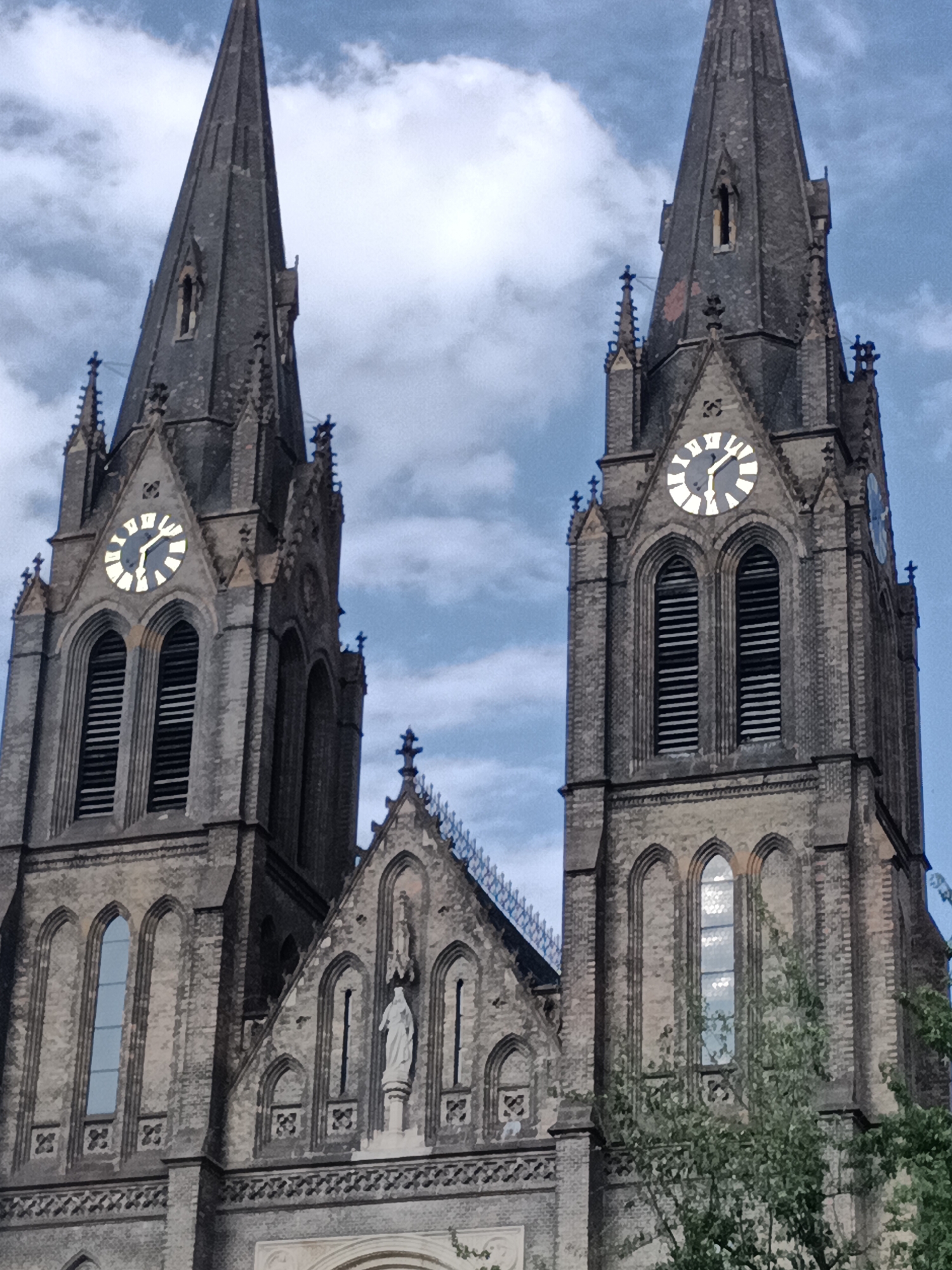 The image depicts the twin spires of the Church of St. Ludmila in Prague, Czechia, located in the Vinohrady district. The church, built in Neo-Gothic style between 1888 and 1892, is characterized by its imposing brick structure and ornate details. The spires, reaching towards a cloudy blue sky, are topped with crosses. Each spire features a large clock face with illuminated Roman numerals, indicating the time. Below the clocks, arched windows with louvers are visible. The facade of the church is adorned with architectural elements, including smaller spires, decorative stonework, and arched doorways. A statue of a female figure, likely St. Ludmila, stands prominently in a niche on the facade. Lush green trees partially obscure the lower section of the church. The overall scene captures a significant architectural landmark against a backdrop of dynamic cloud formations.