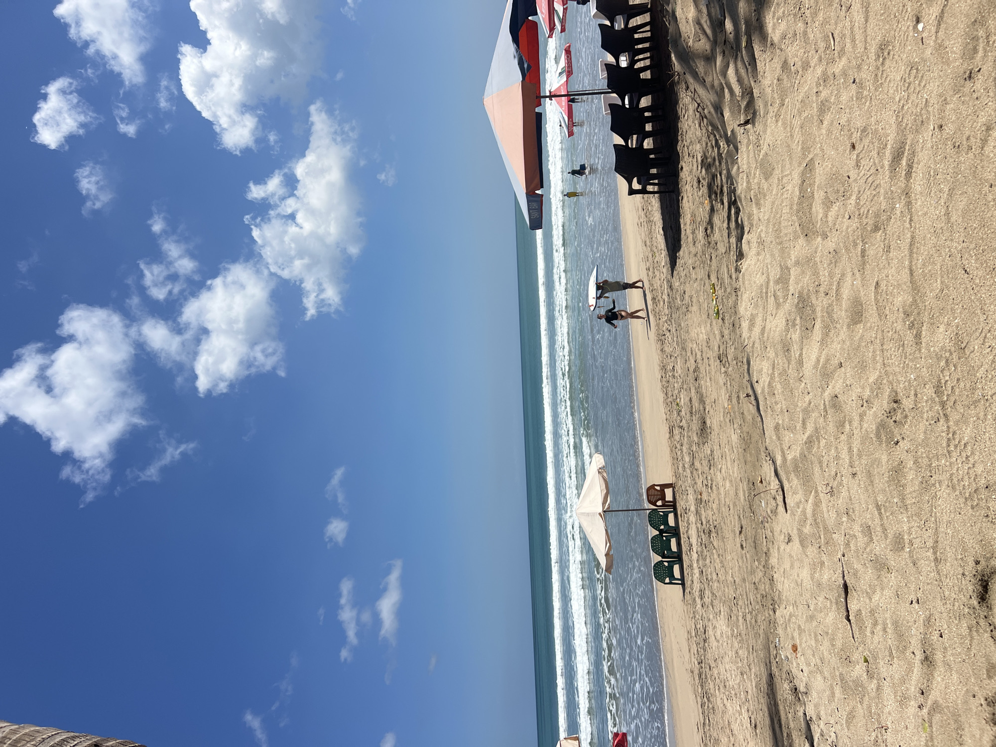On a sunny day at Kuta Beach, Bali, Indonesia, the sandy shore is dotted with beach chairs and umbrellas. The ocean waves roll onto the shore under a blue sky with scattered white clouds. Two people are walking near the water's edge, one carrying a surfboard. In the background, other beachgoers are enjoying the water. The scene captures a typical day at this popular Indonesian beach destination.